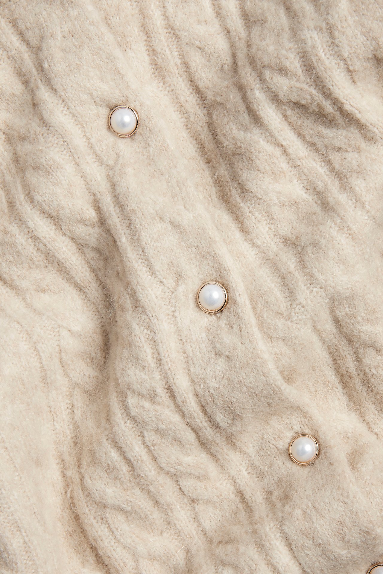 Nærbilde av en beige kabelstrikket cardigan med runde perleknapper og gullfargede kanter.