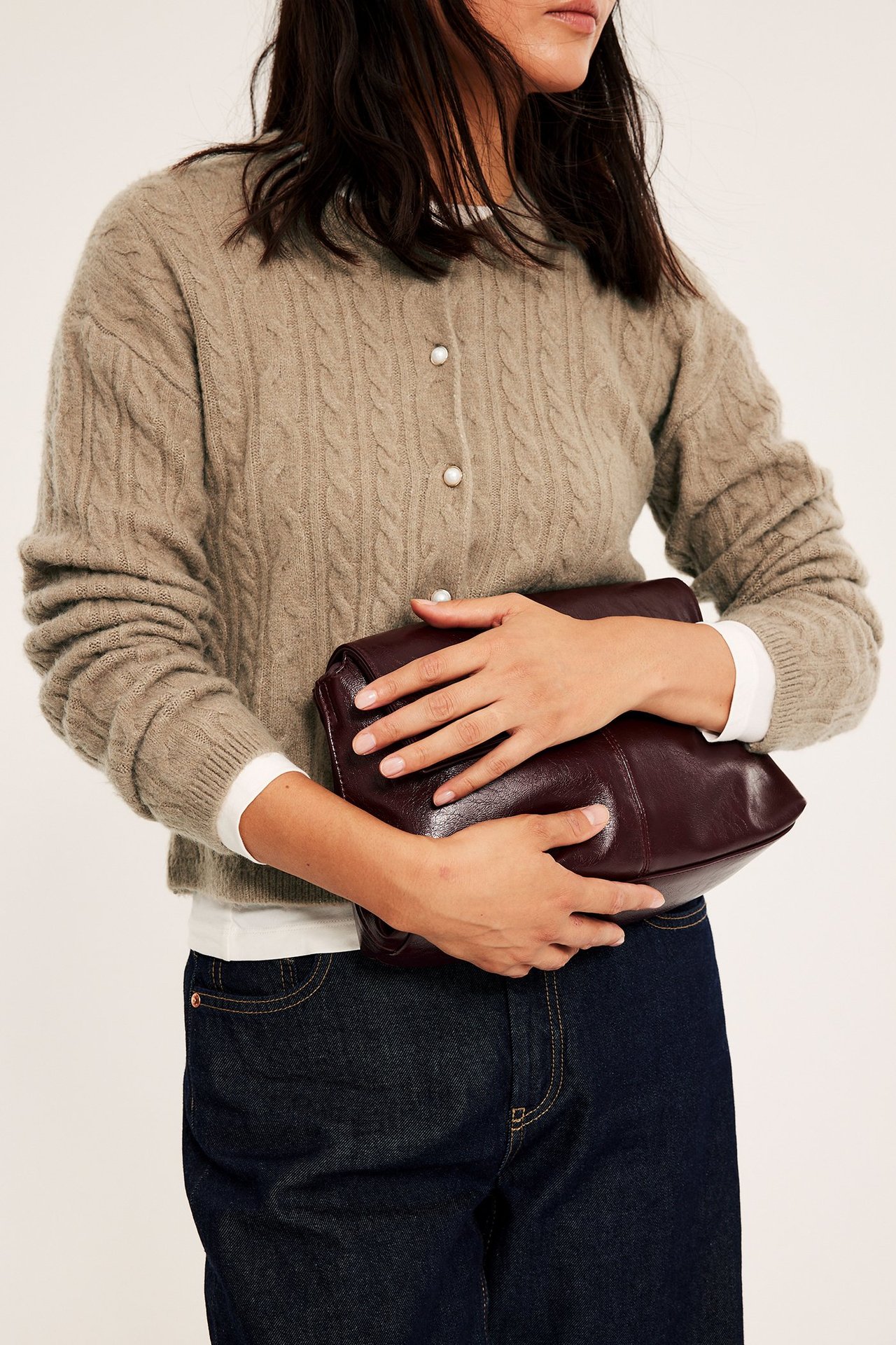 Kobieta nosi ciemnobeżowy sweter z splotem i perłowymi guzami, trzyma ciemnoczerwoną torbę.