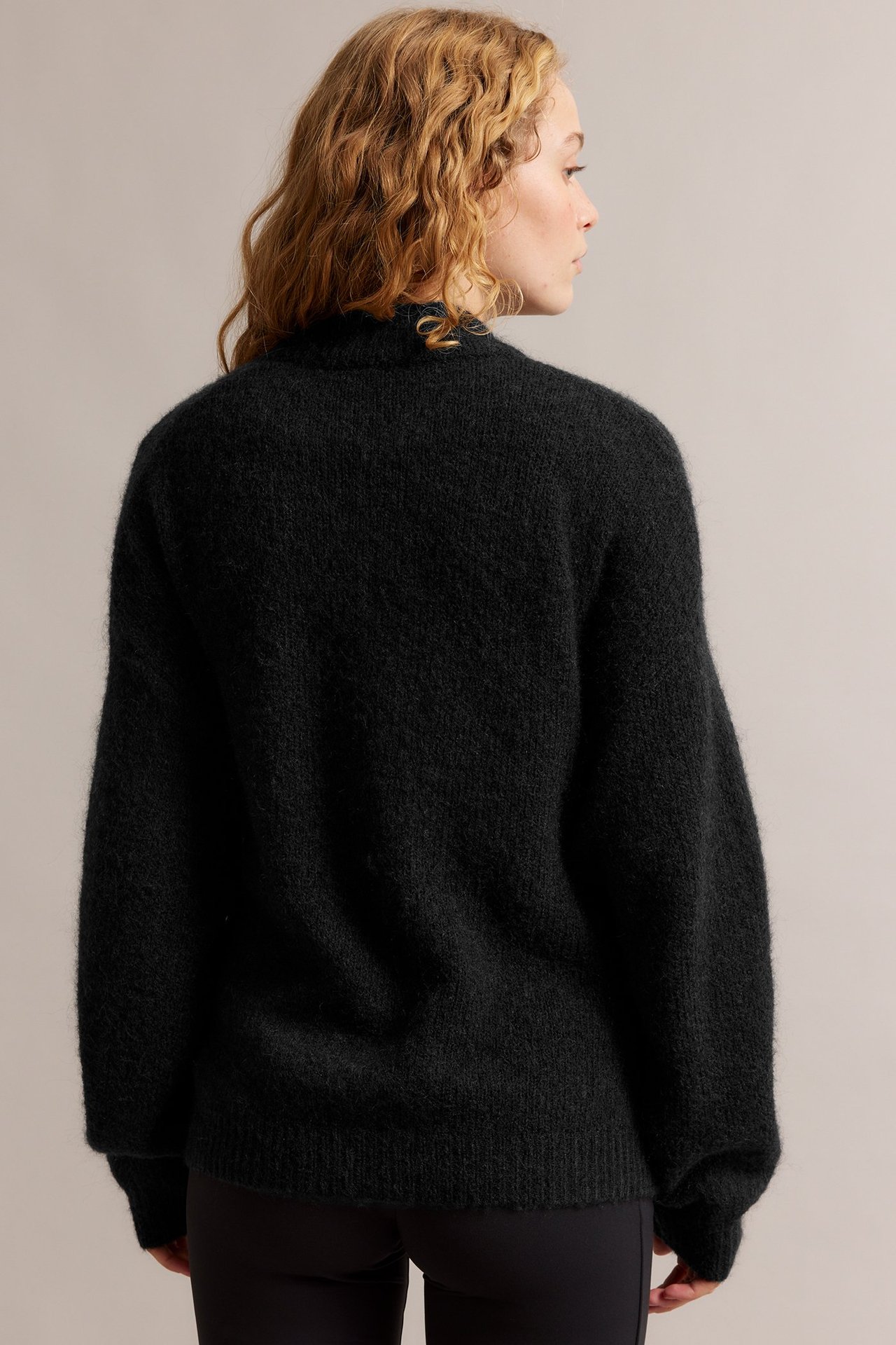 Naisen takanäkymä yllään musta, löysä villasekoitekardigaani ja mustat housut.