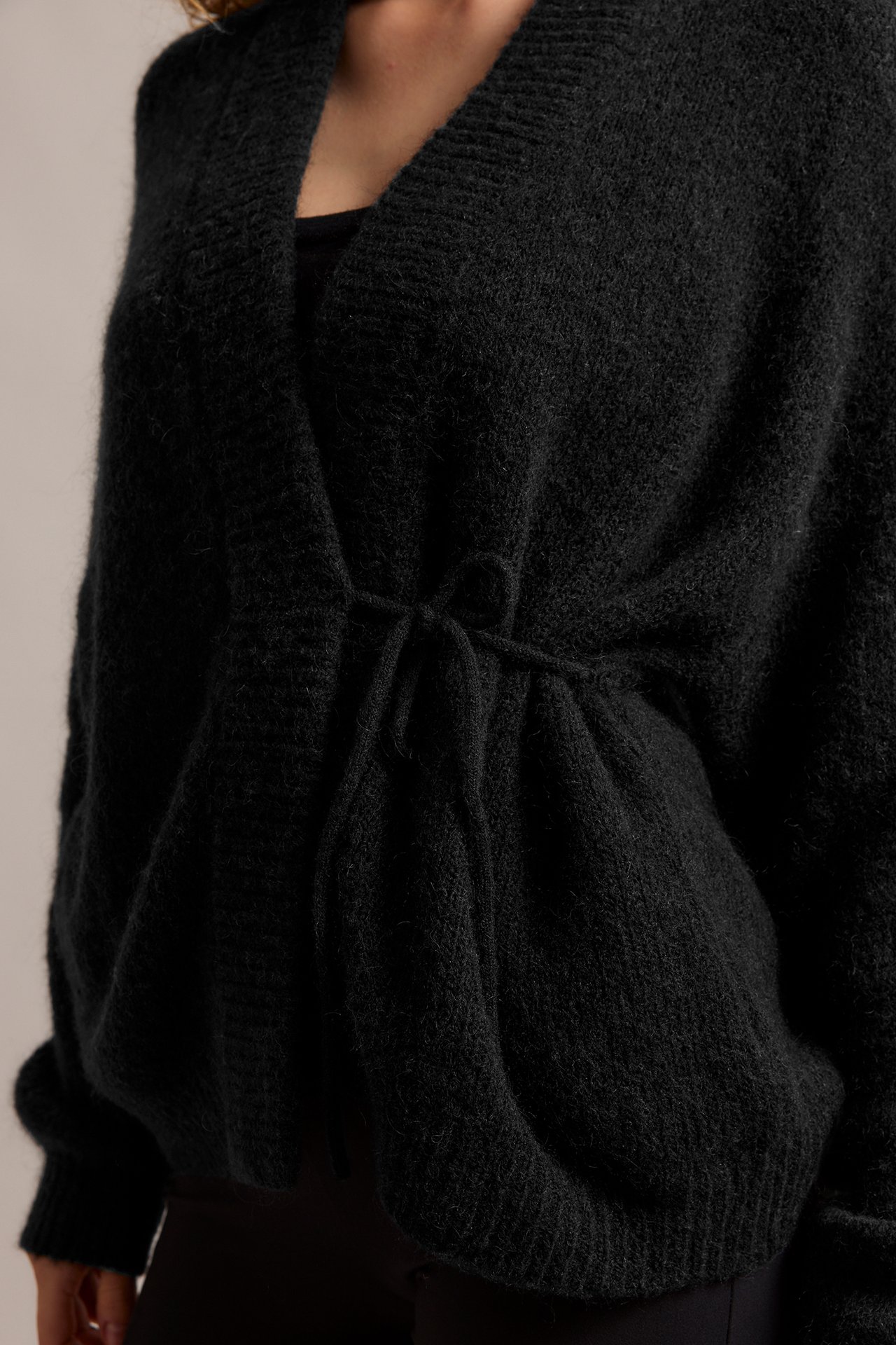 Lähikuva edestä: nainen yllään musta villasekoite-kietaisuneuletakki solmittavalla nauhalla tumman topin päällä.