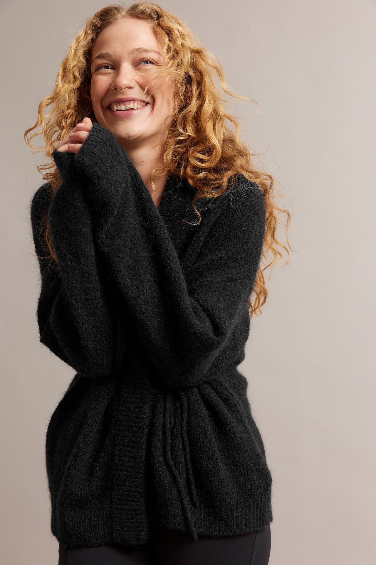 Forfra, smilende kvinne i en svart ullblandingskardigan, knyttet i midjen.