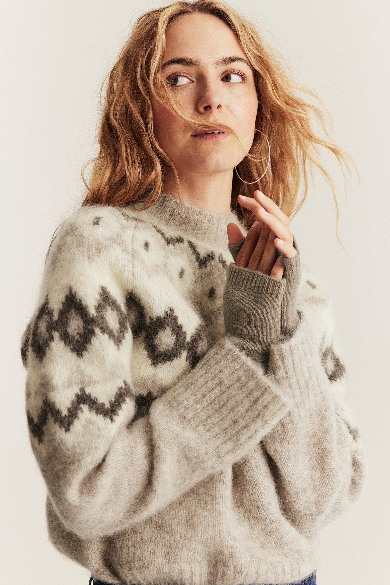 Lähikuva naisesta, jolla on beigemelange-villa-kashmir-rannekkeet kuvioidun harmaan neuleen päällä ja kultaiset korvarenkaat.