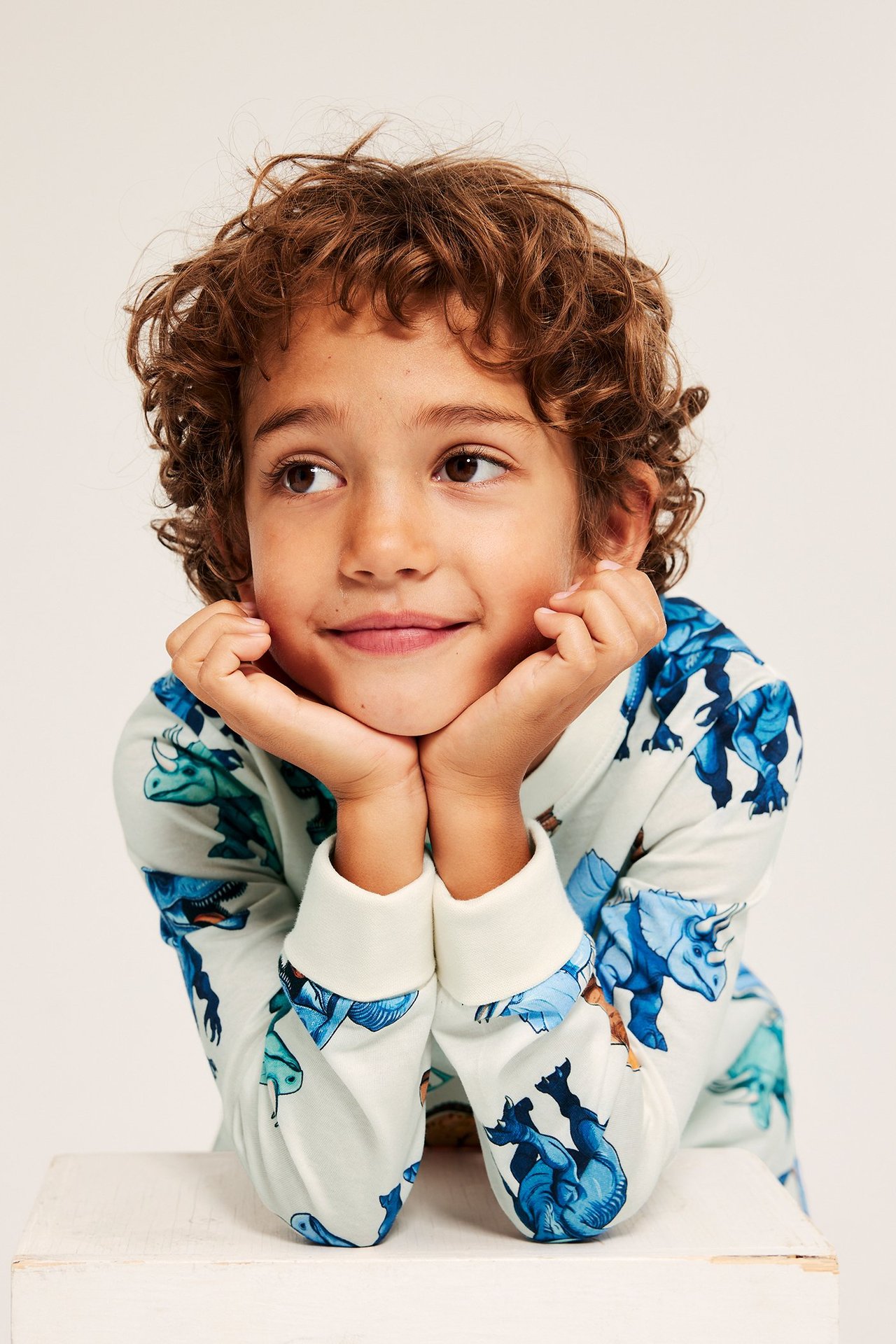 Lähikuva lapsesta yllään luonnonvalkoinen puuvillapyjamatoppi sinisellä dinosauruskuviolla ja resoreilla, kädet leuan alla.