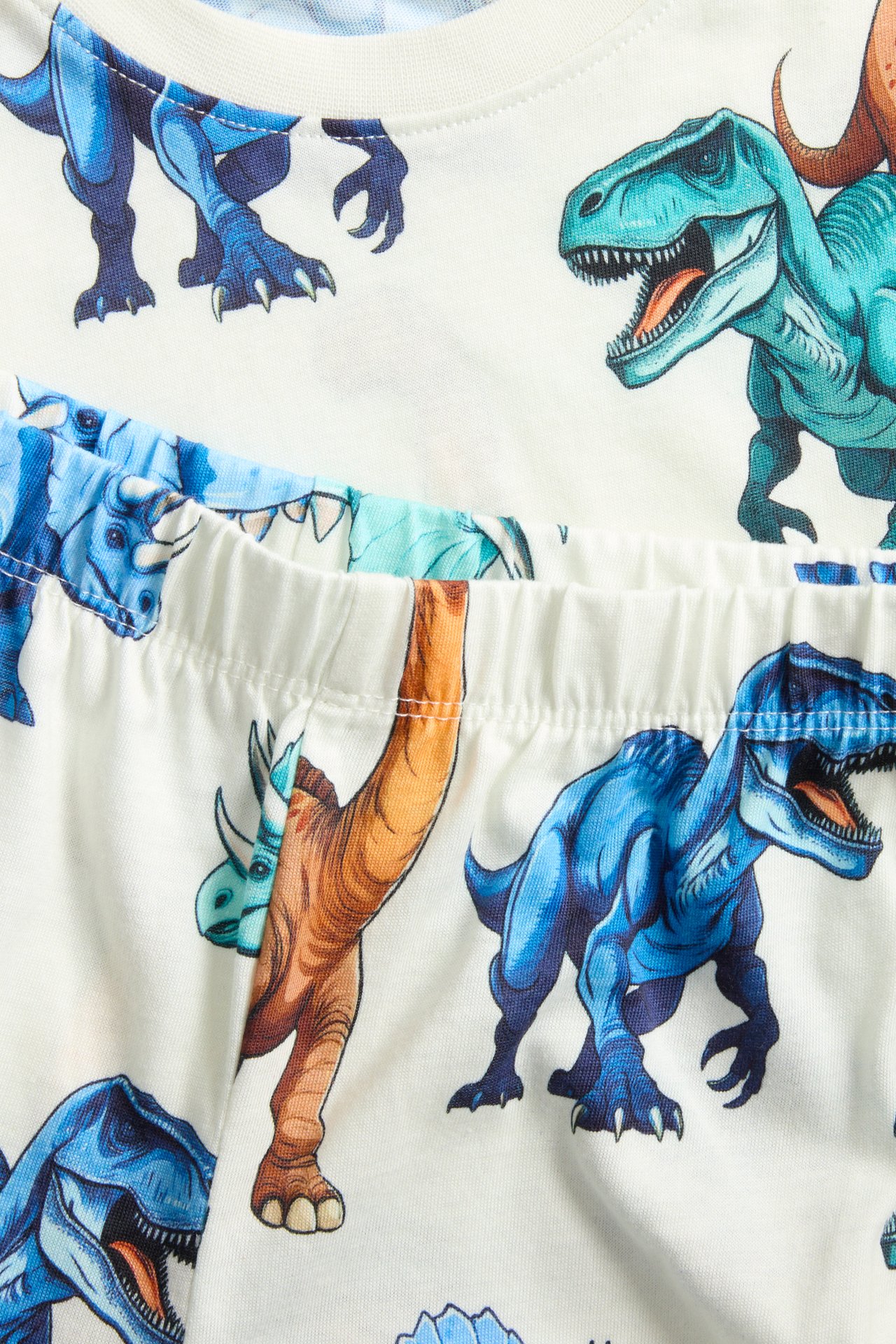 Lähikuva luonnonvalkoisesta puuvillapyjamasetistä lapsille, sinisillä ja oransseilla dinosauruskuvioilla.