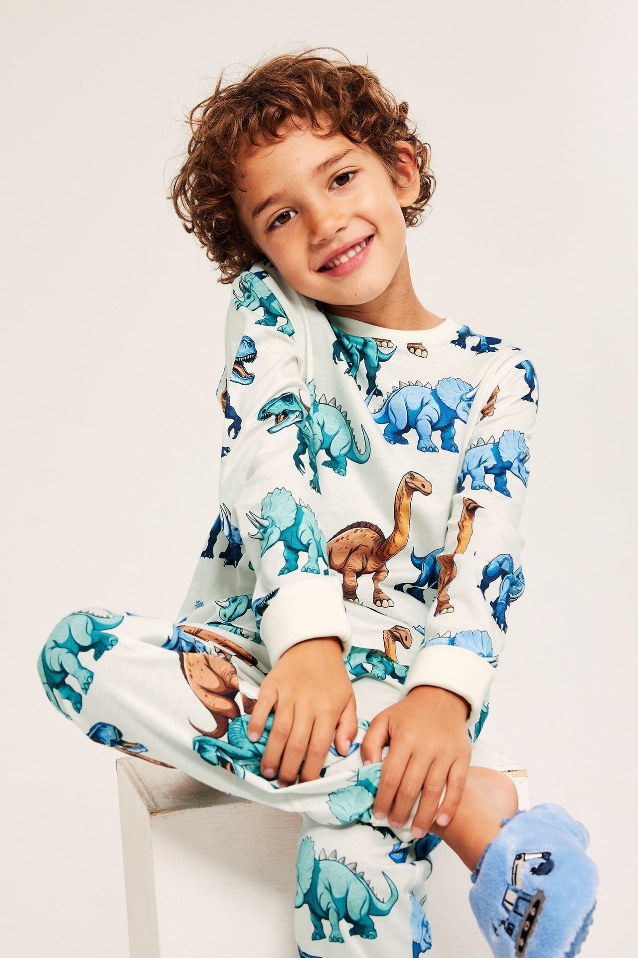 Edestäpäin: lapsi yllään luonnonvalkoiset puuvillapyjamat värikkäällä dinosauruskuviolla ja siniset tossut.