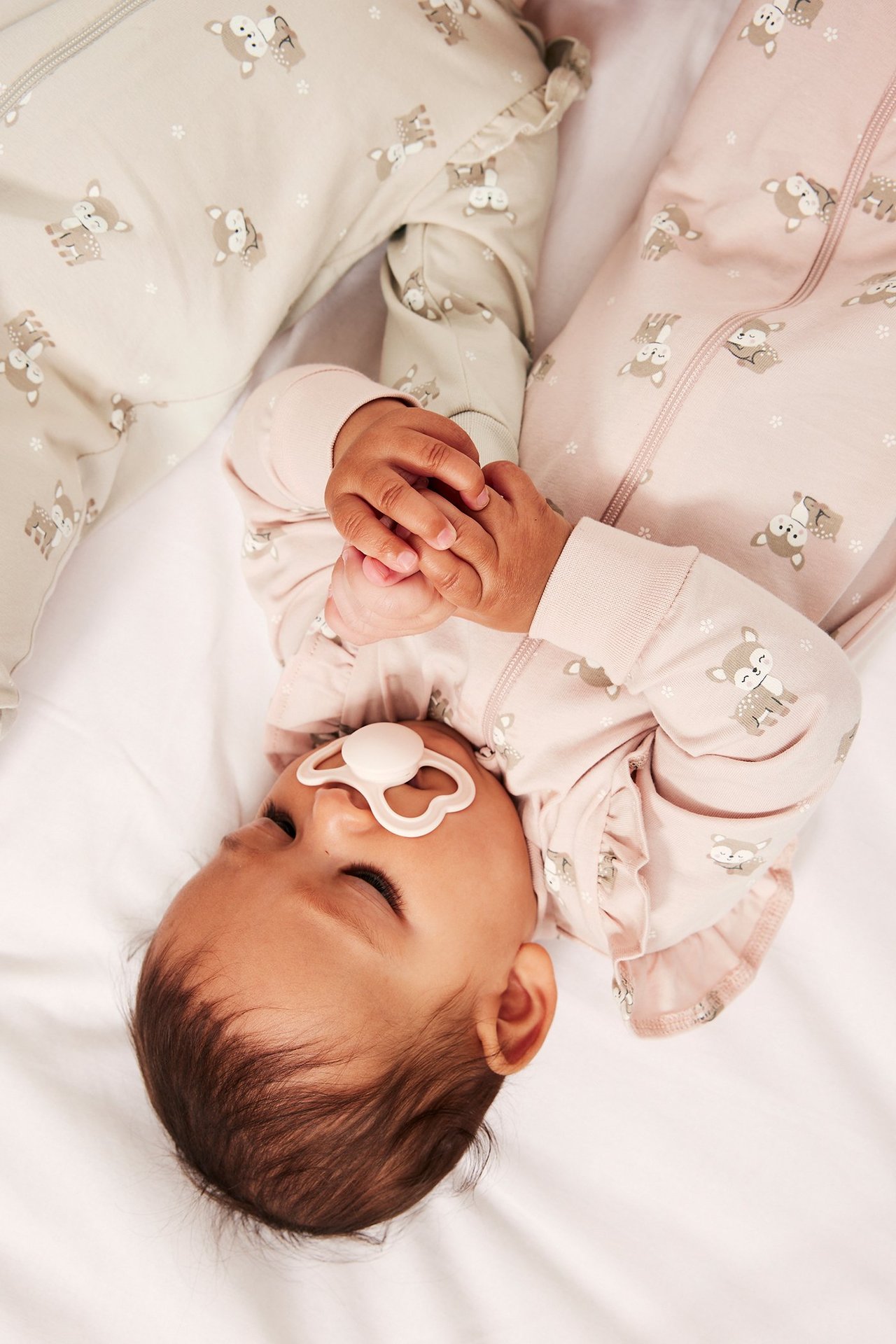 Baby i rosa rådjursmönstrad pyjamas med volanger, dragkedja, håller vit napp. Beige pyjamas syns.