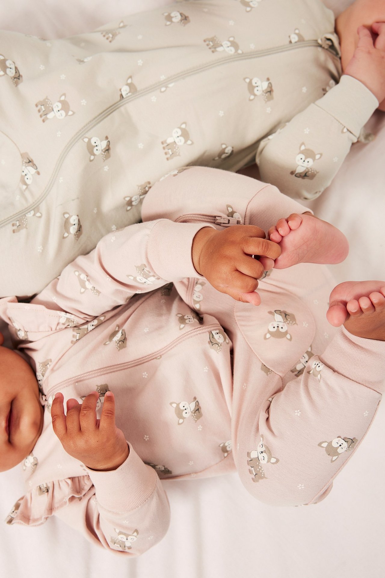 Två bebisar bär mönstrade pyjamasar: en rosa med volanger och dragkedja, den andra beige med dragkedja.
