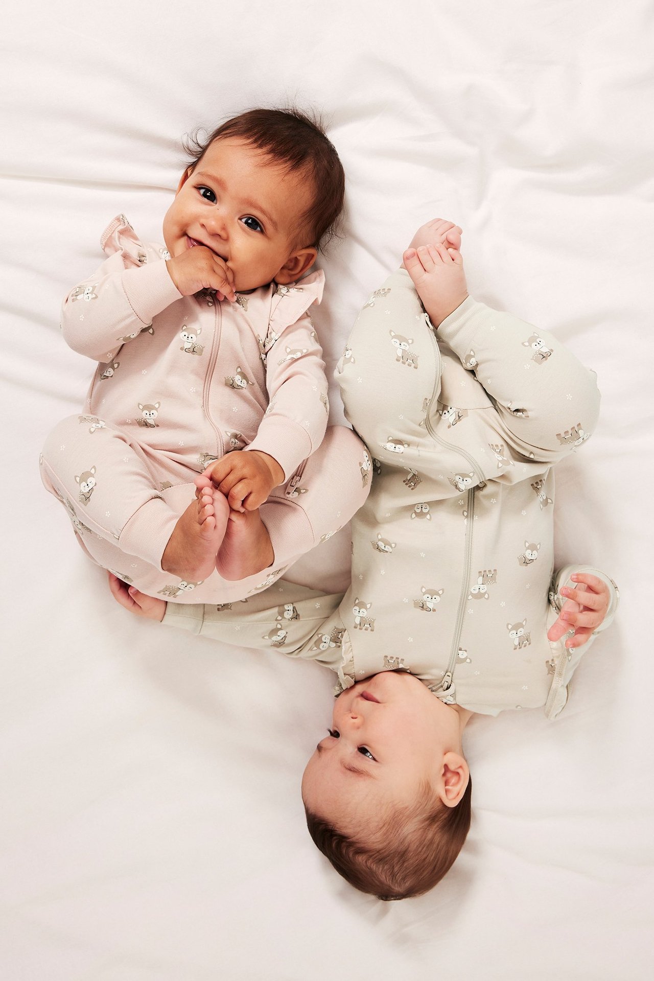 Uppifrån-vy av två bebisar i mönstrade bomullsrompers. En bär rosa volangromper, den andra en beige.