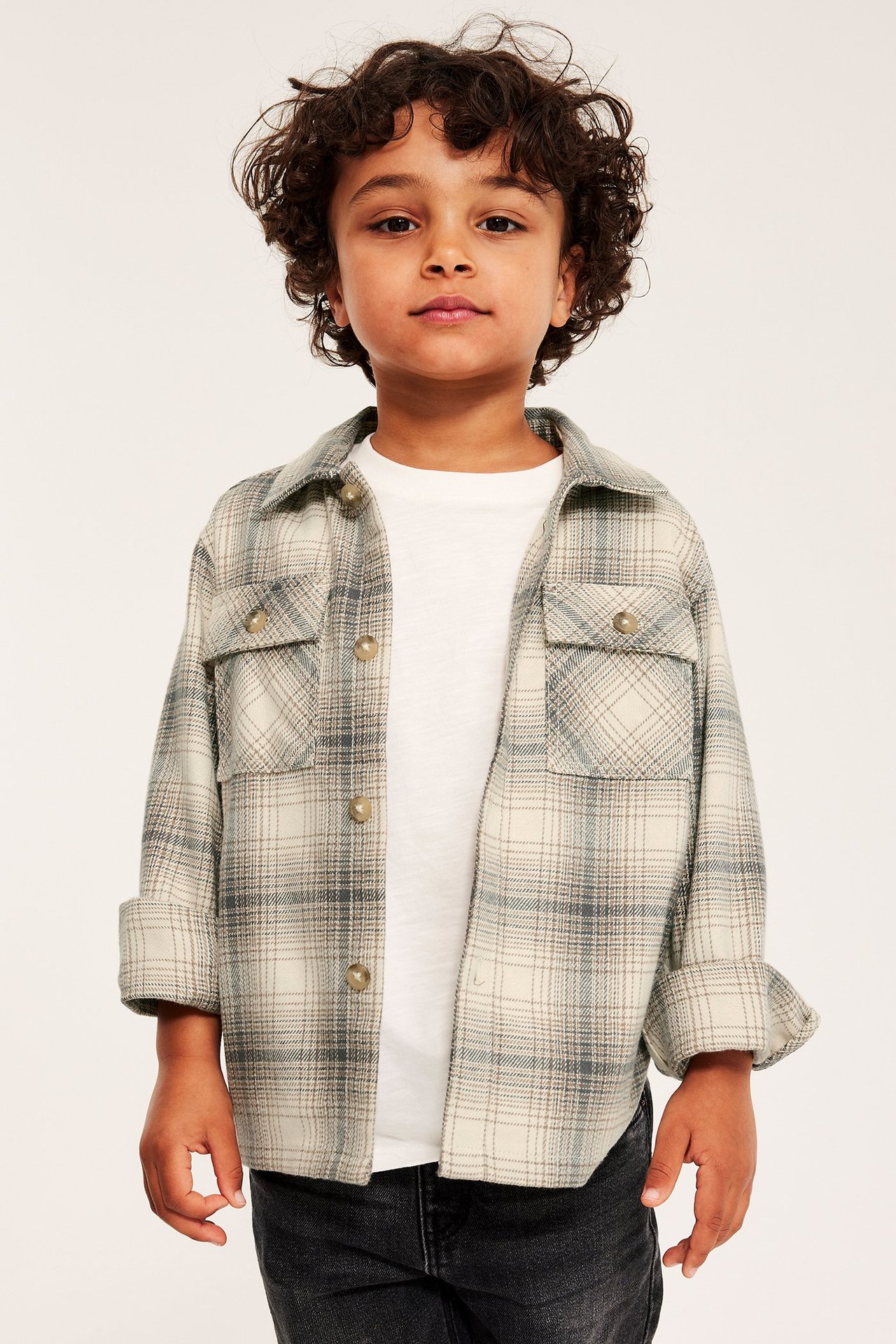 Barnkille i rutig, ljusbeige overshirt i flanell, med bröstfickor och knappar, över vit t-shirt.