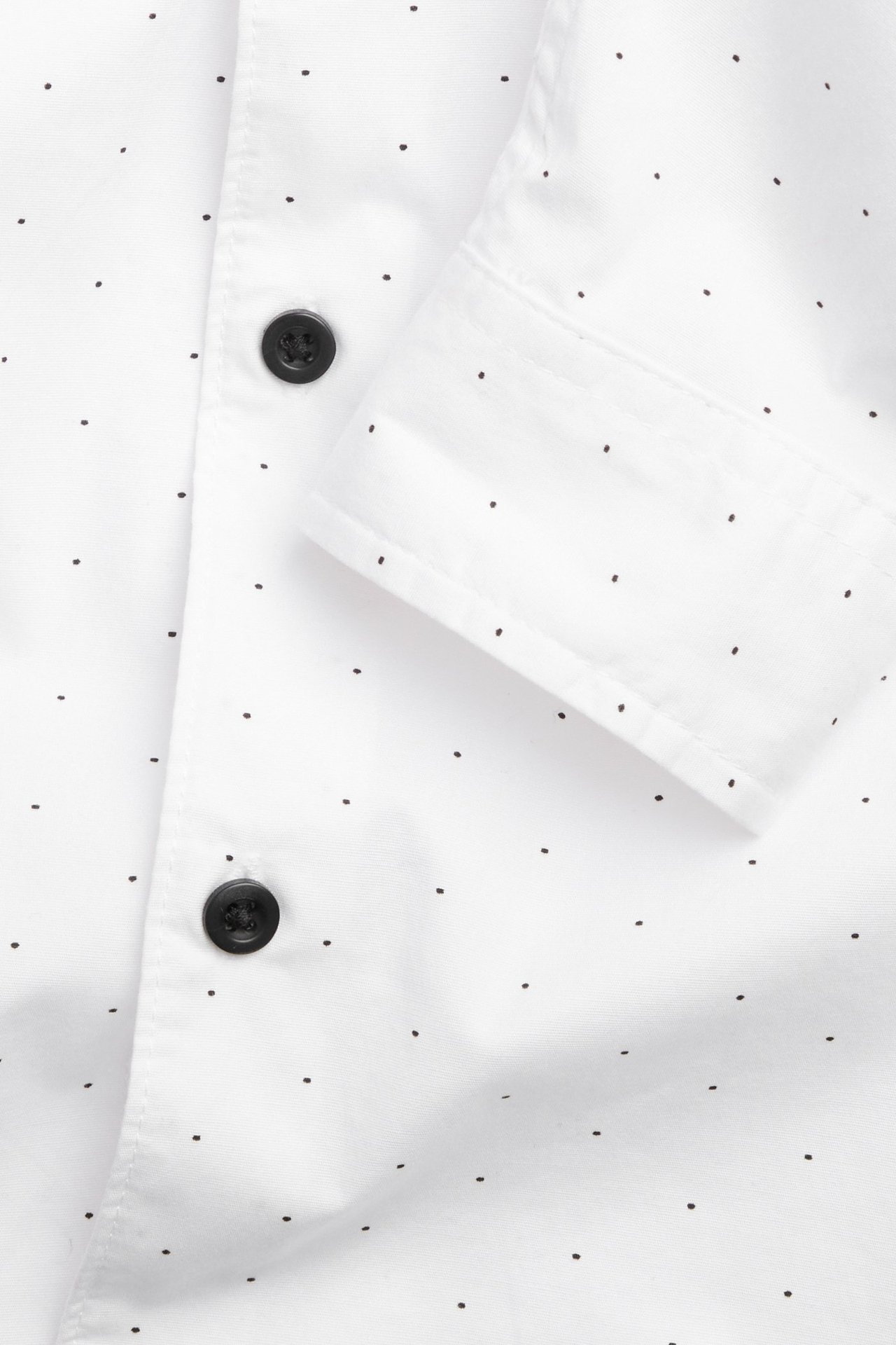 Zbliżenie białej bawełnianej koszuli dziecięcej w kropki, z czarnymi guzikami i mankietem.