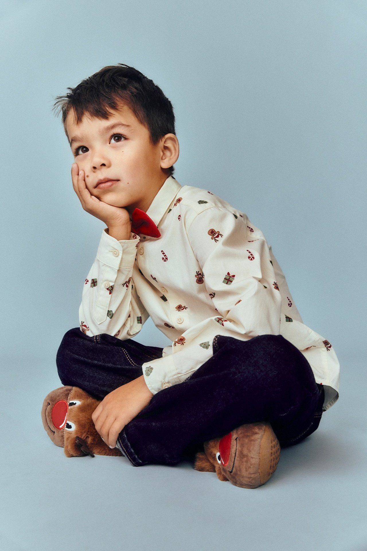 Widok z przodu: Dziecko w jasnobeżowej wzorzystej koszuli bawełnianej, ciemnoniebieskich jeansach, czerwonej muszce i kapciach renifery.