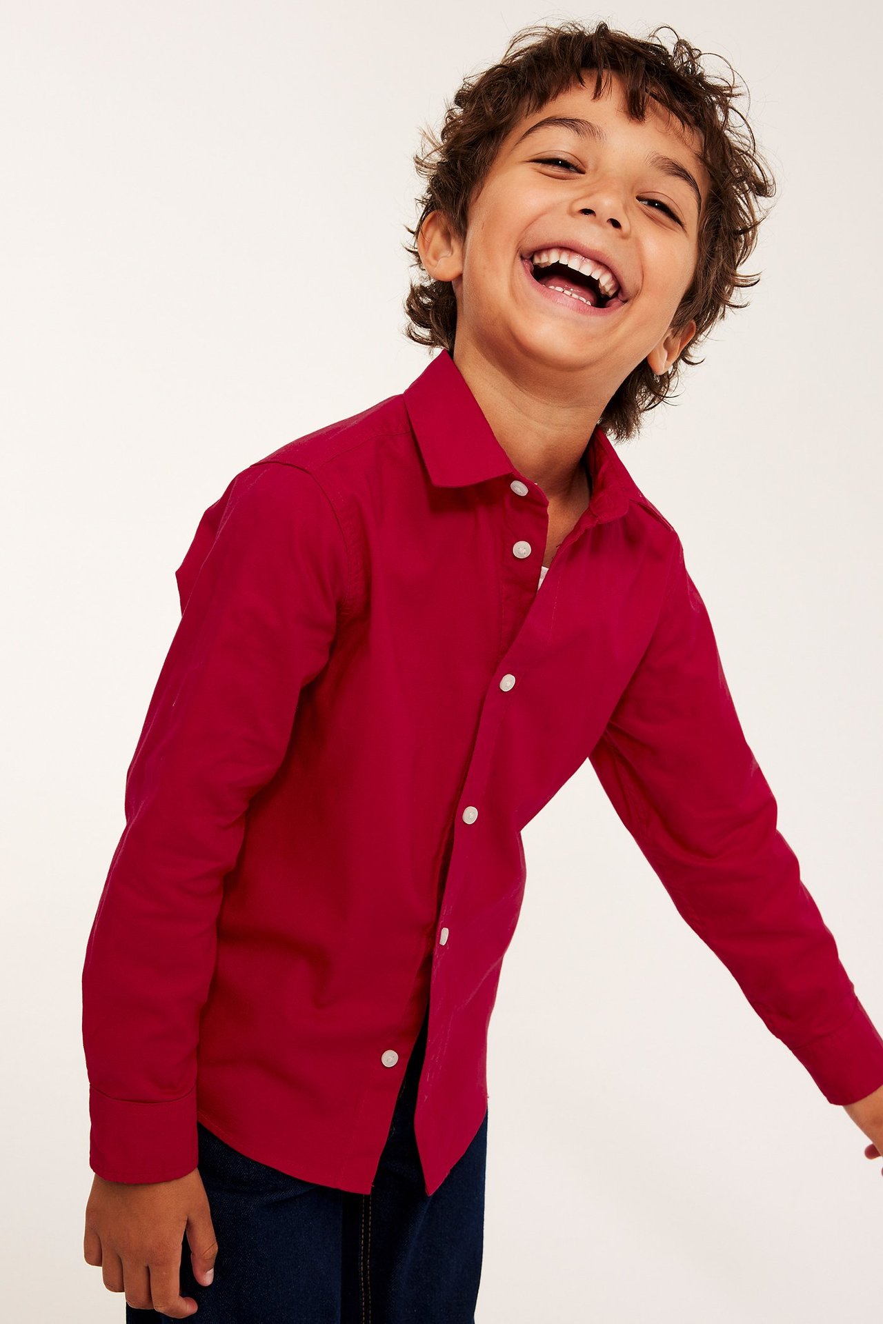 Frontvy av ett skrattande barn i en röd bomulls-oxfordskjorta med vita knappar och mörkblå jeans.