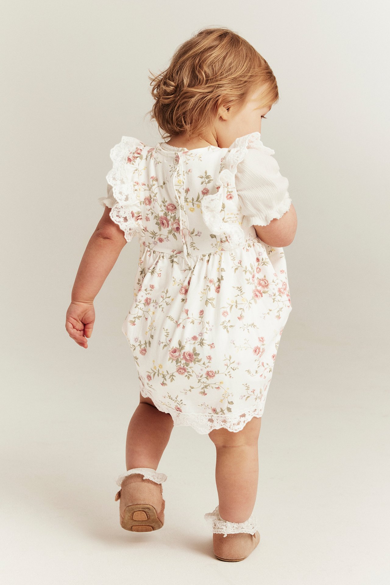 Bakfra: Baby i offwhite blomsterkjole uten ermer med blonder, over hvit topp, brune sko.
