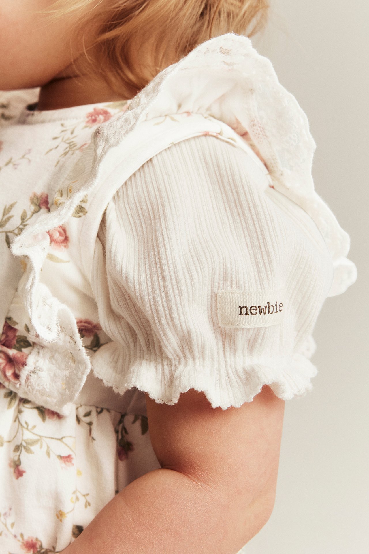 Nærbilde av baby i offwhite blomstret ermeløs kjole med blonder over en ribbet hvit topp.