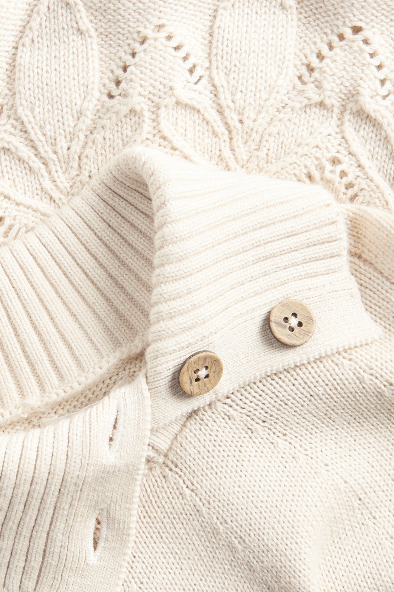 Zbliżenie beżowego dzianinowego poncza bawełnianego ze wzorem warkoczowym i drewnianymi guzikami.