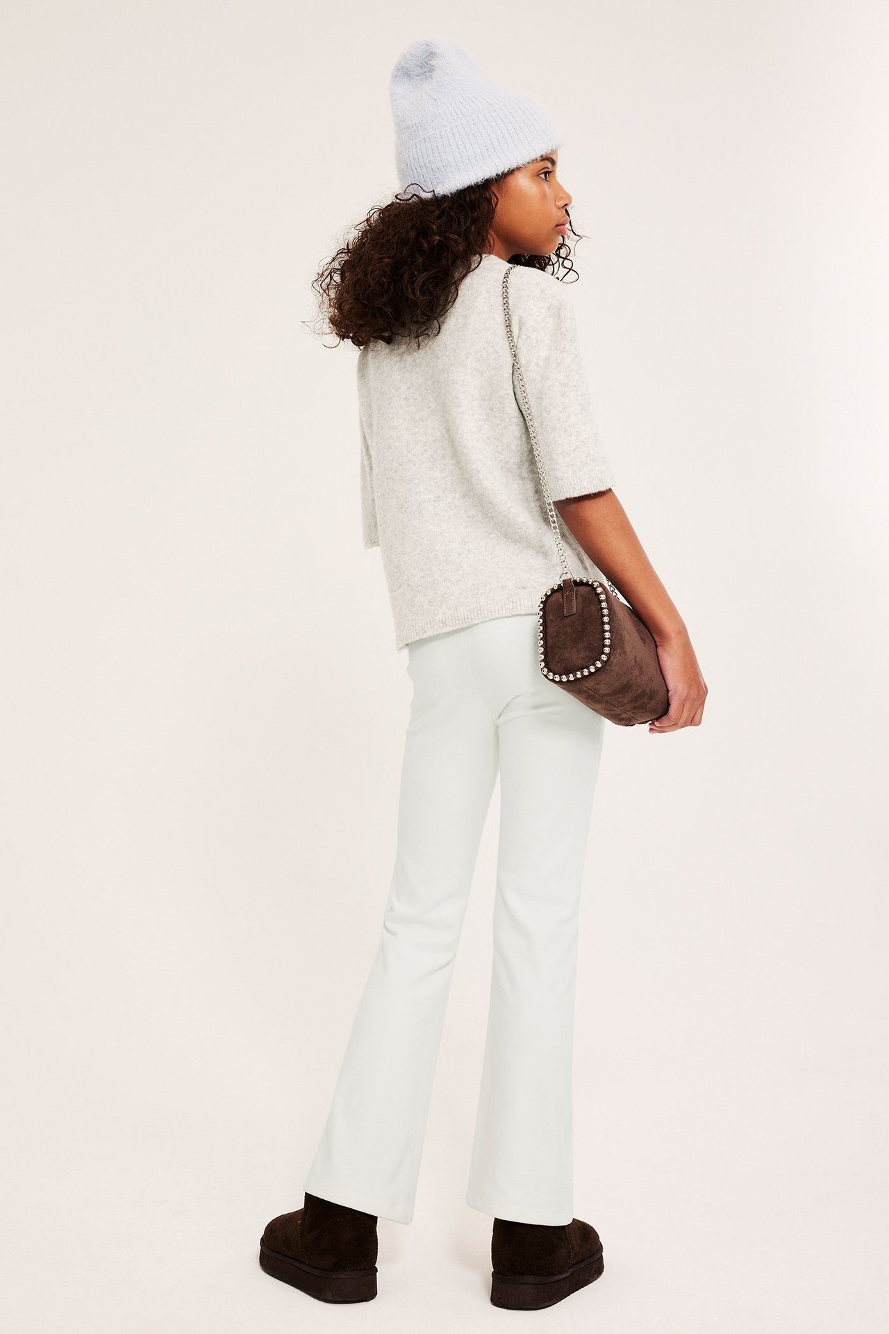 Sned bakifrån: Barn i offwhite utsvängda yogabyxor, ljusgrå topp, ljusblå mössa, brun väska, bruna kängor.