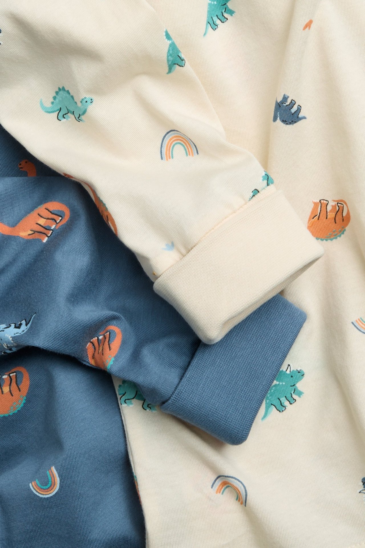 Zbliżenie dwóch bawełnianych bluzek piżamowych dla dzieci, beżowej i niebieskiej, z nadrukiem dinozaurów i mankietami.
