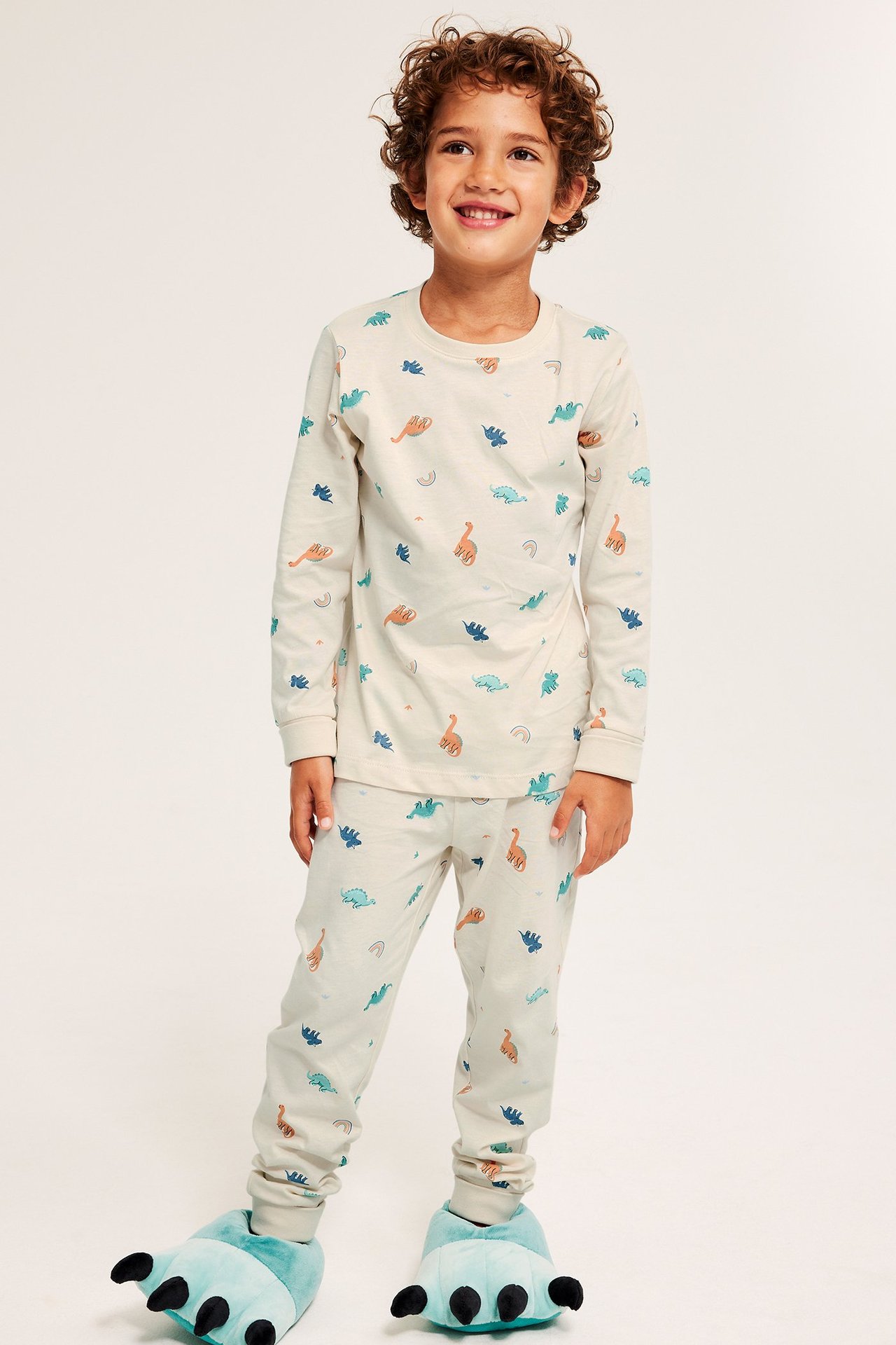 Edestäpäin: Lapsi yllään vaalea beige puuvillapyjamas dinosauruskuviolla ja siniset hirviötossut.