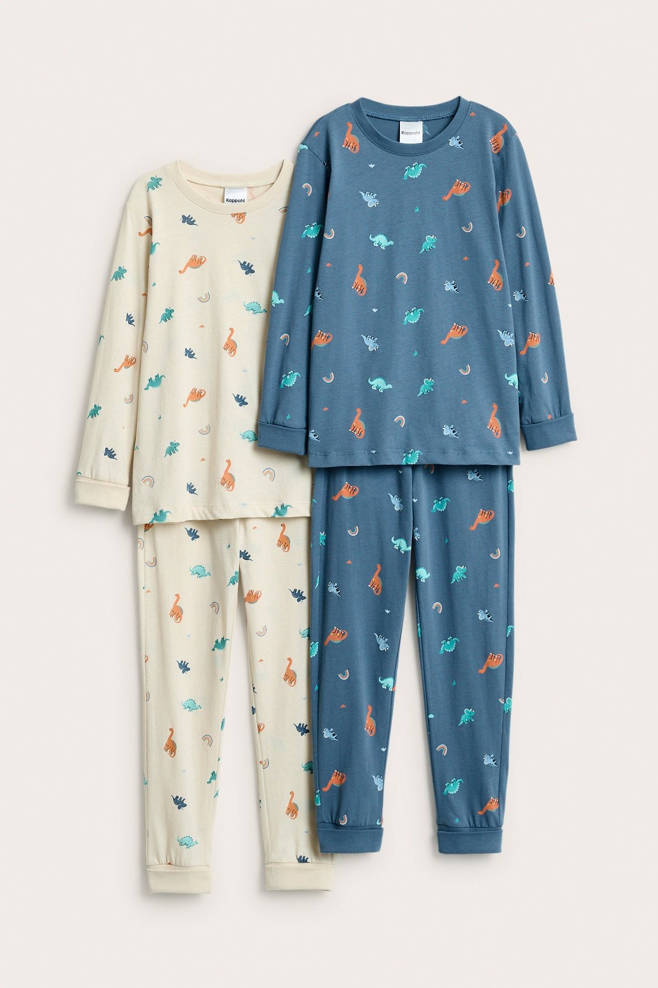 Edestä kahden lasten pyjamas-setin näkymä: toinen vaalea beige, toinen sininen, molemmissa dinoprintti.