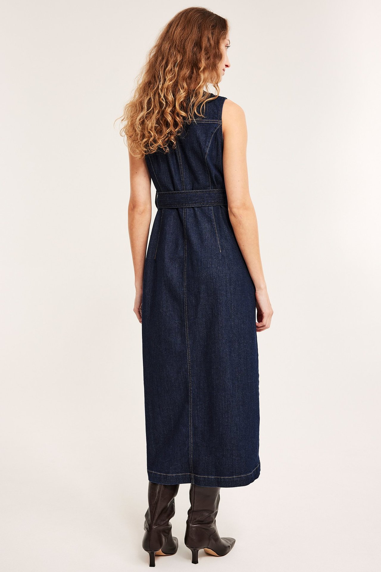 Ryggvy av kvinna i mörkblå ärmlös midilång jeansskjortklänning med bälte, bärande mörkbruna stövlar.