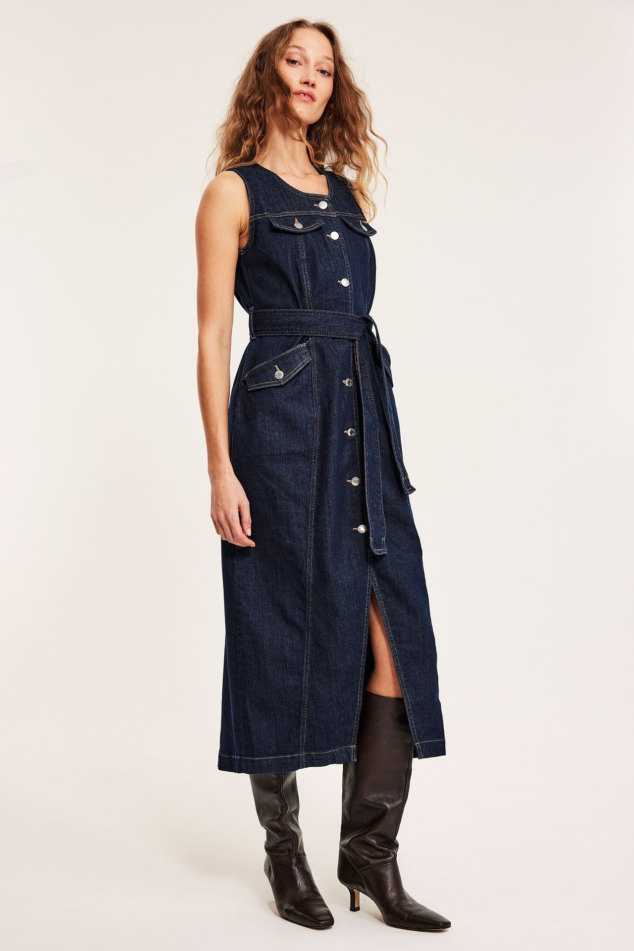Framifrån: kvinna i mörk ärmlös jeansklänning med silverknappar och knytband i midjan, stylad med bruna stövlar.