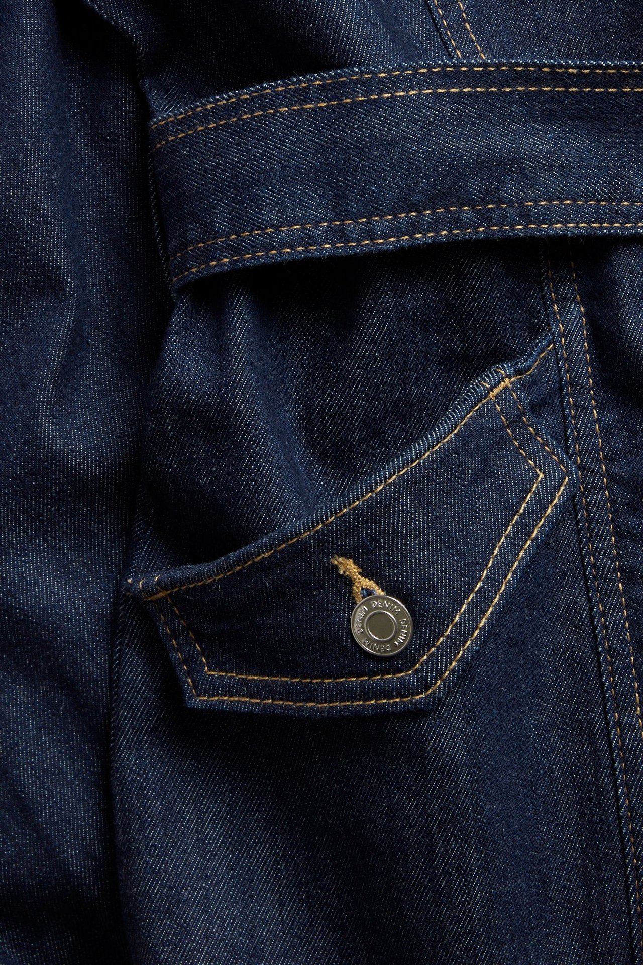 Närbild på en mörk jeansklänning med ljusa kontrastsömmar, en ficklock och en silverfärgad knapp.