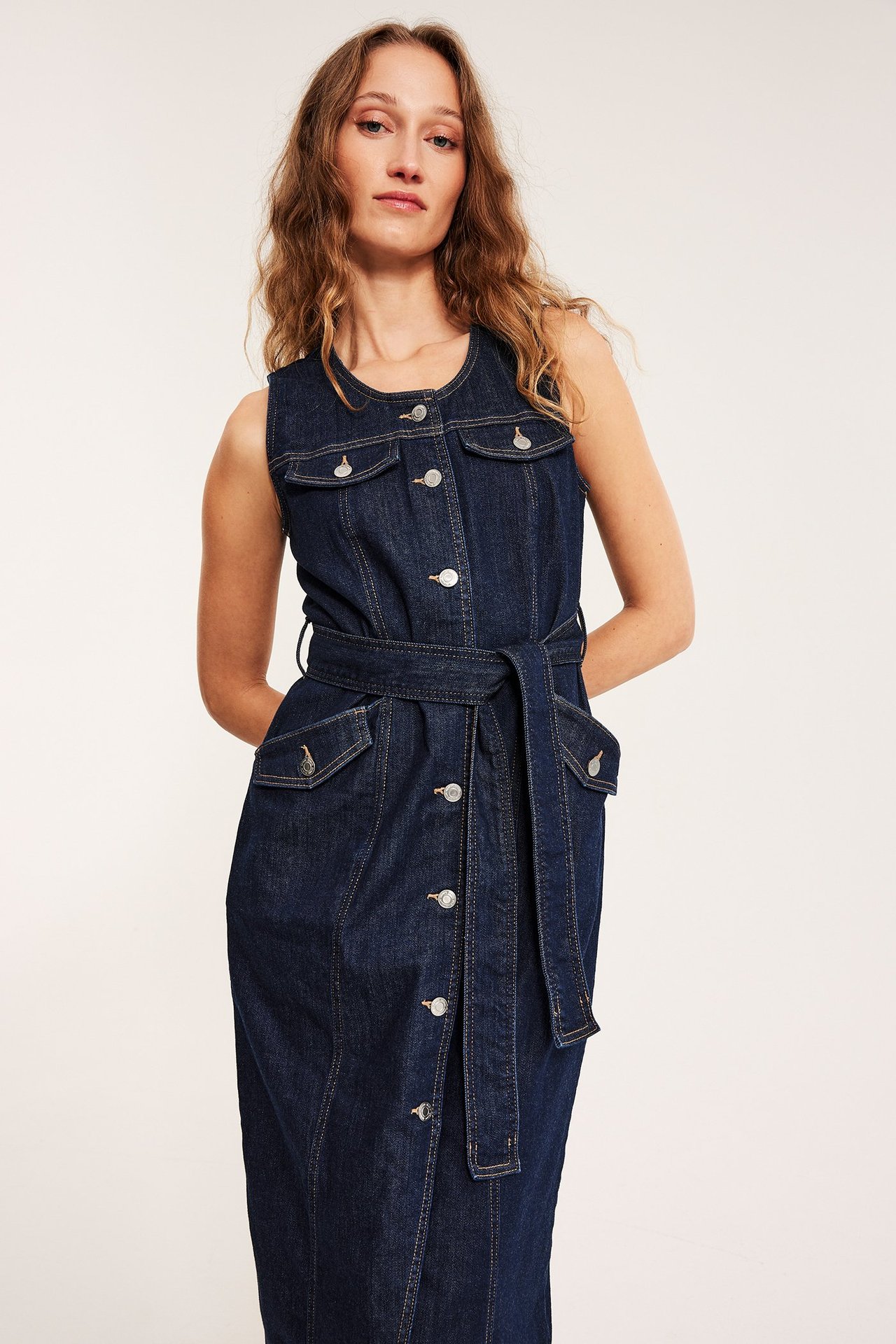 Framifrån: kvinna i mörk jeansblå ärmlös midiklänning med silverknappar och knytband i midjan.