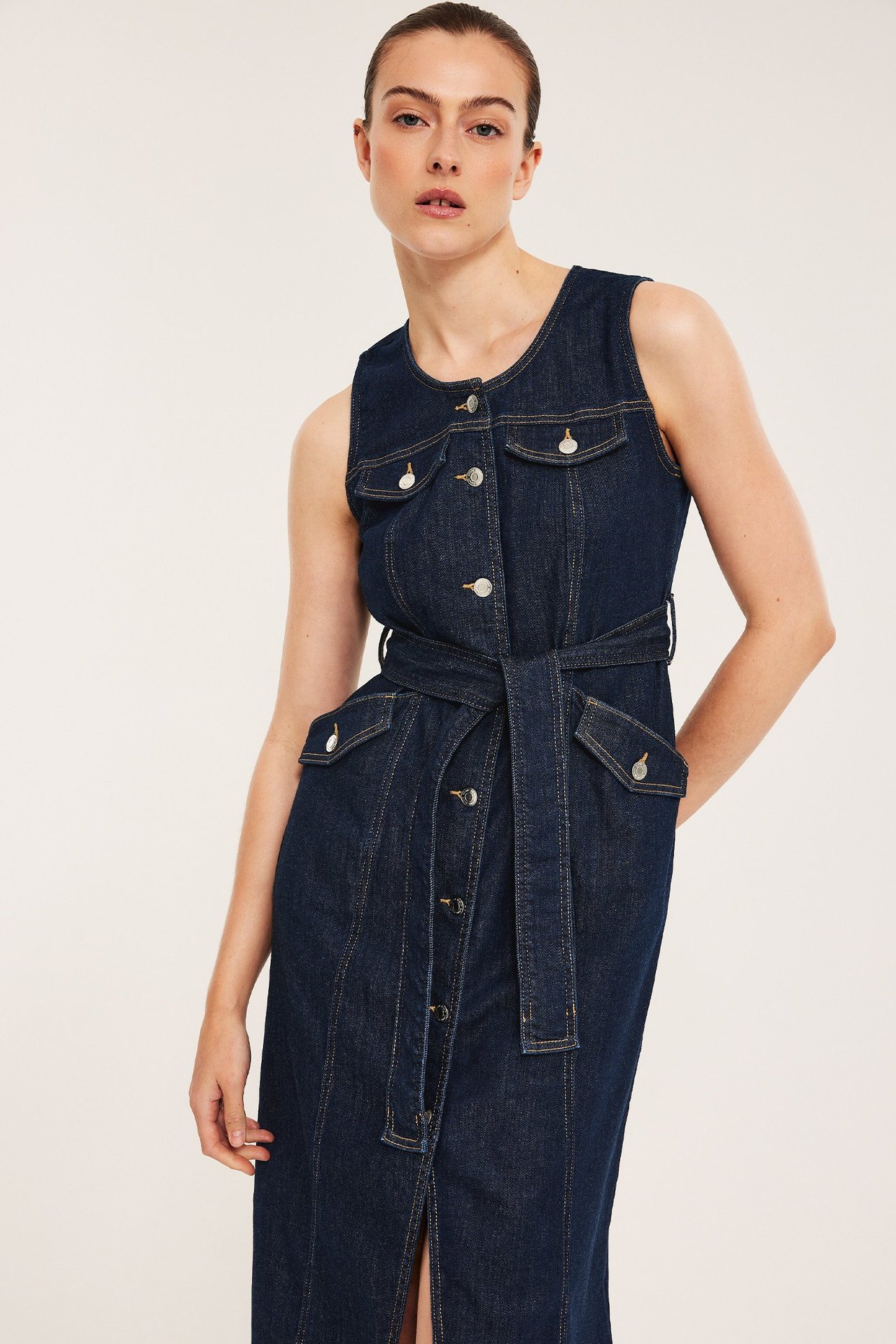 Frontvisning: kvinne i mørk, ermeløs denim midikjole med knapper, klafflommer og belte.