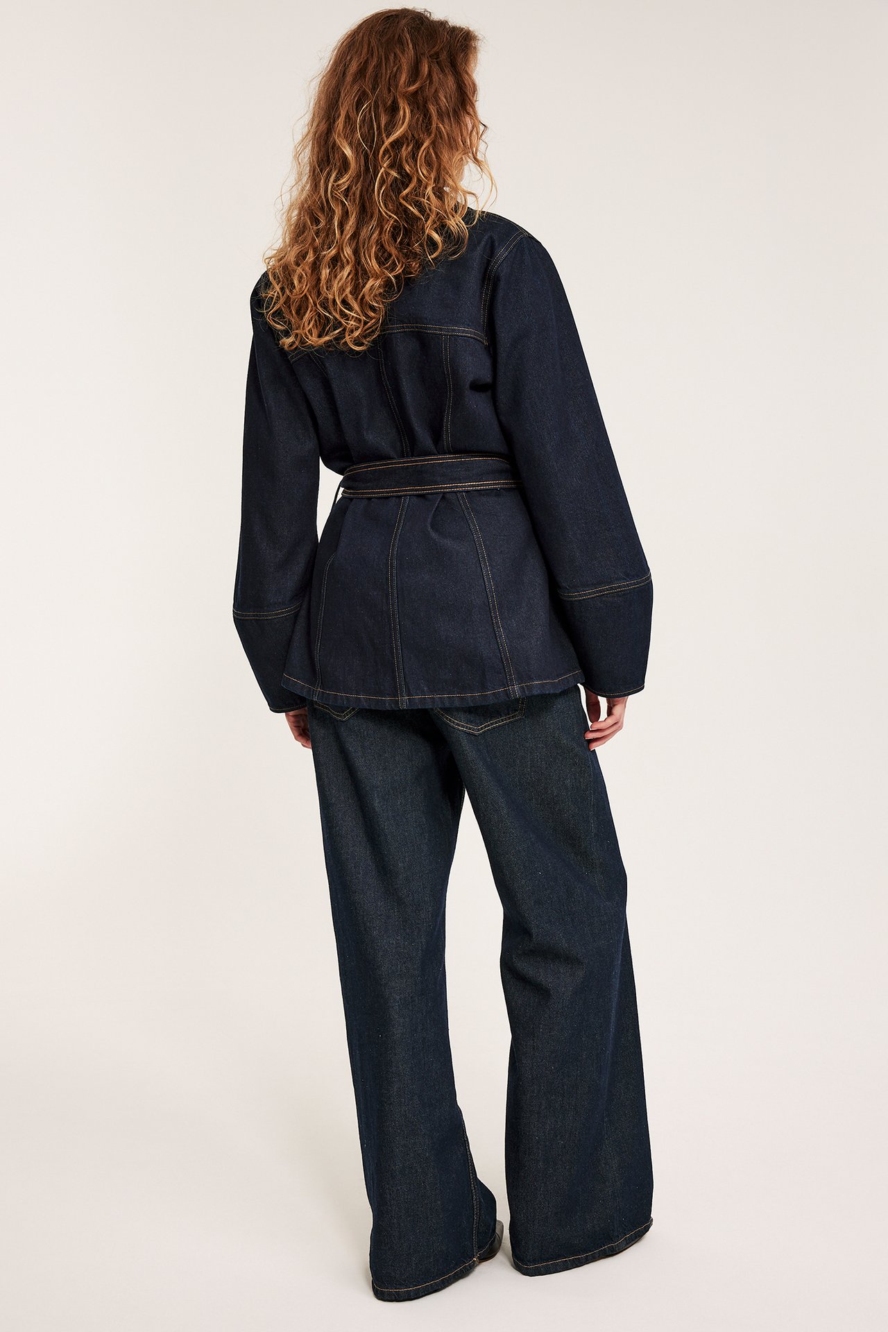 Naisen takaa näkymä tummansinisestä farkkutakista vyöllä ja yhteensopivista leveälahkeisista farkuista.