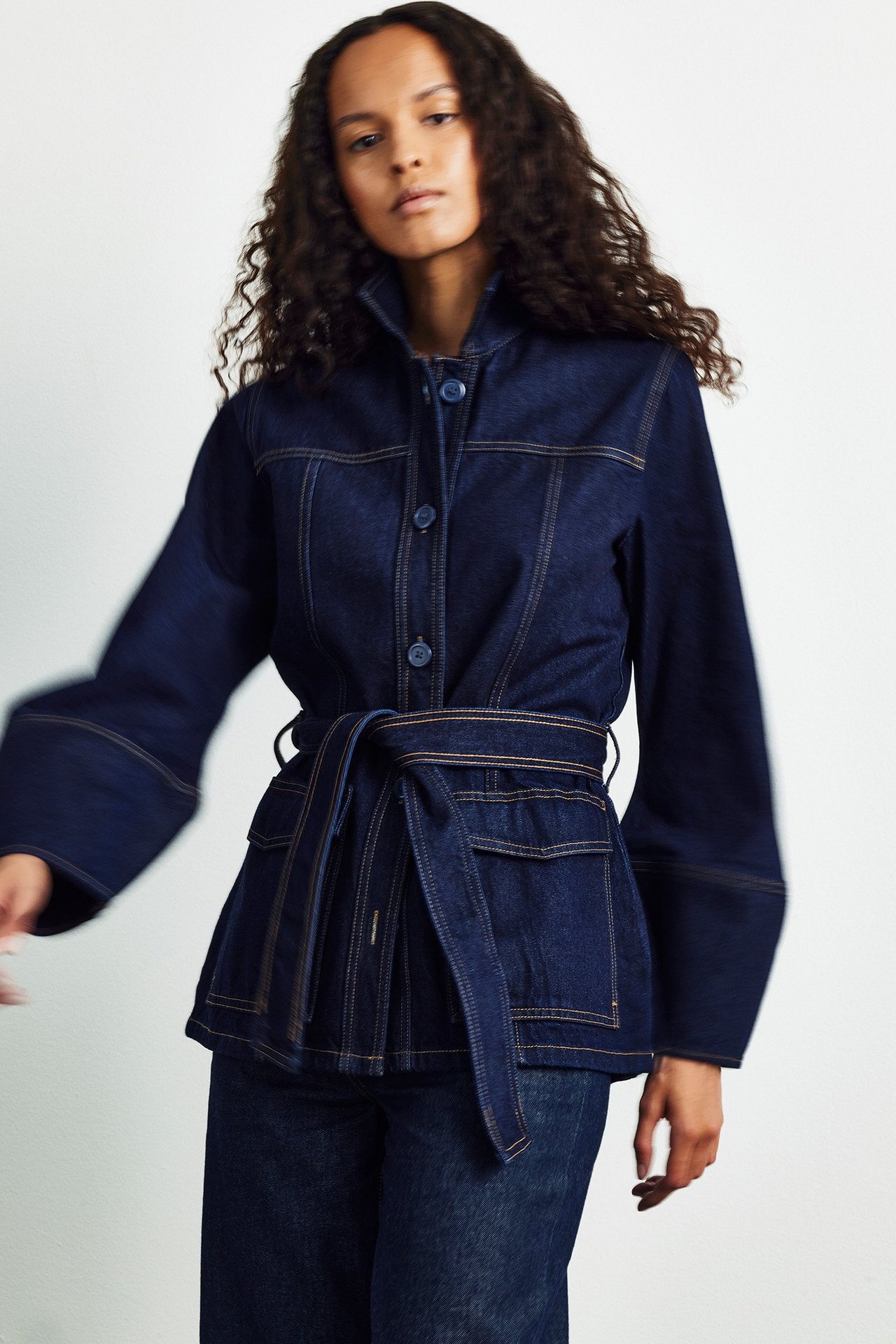 Nainen tummansinisessä farkkutakissa läpällisillä taskuilla, solmitulla vyöllä ja tummissa farkuissa, edestä.