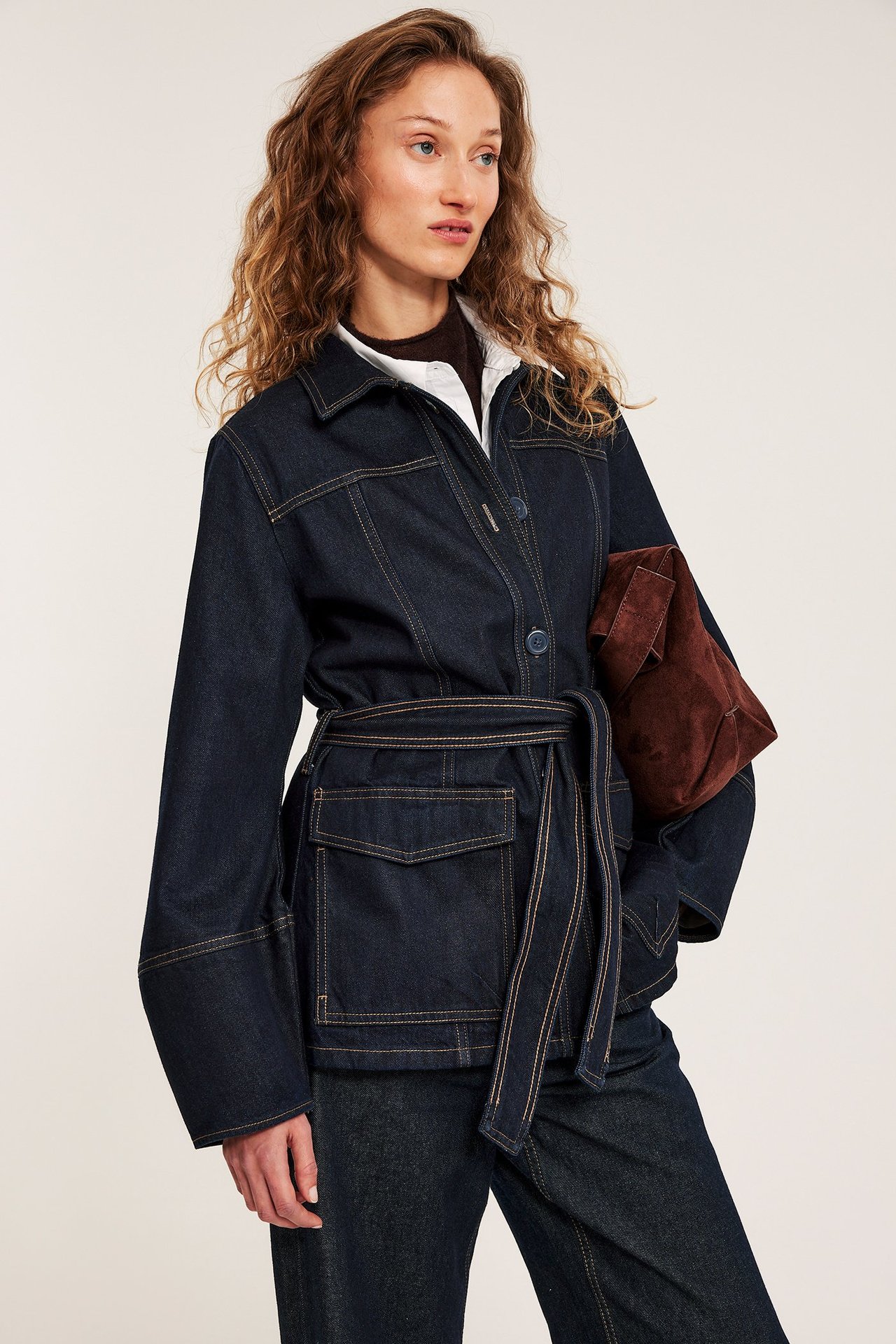 Edestäpäin: nainen tummansinisessä vyöllisessä farkkutakissa, tummissa housuissa, pitäen ruskeaa mokkakytkintä.