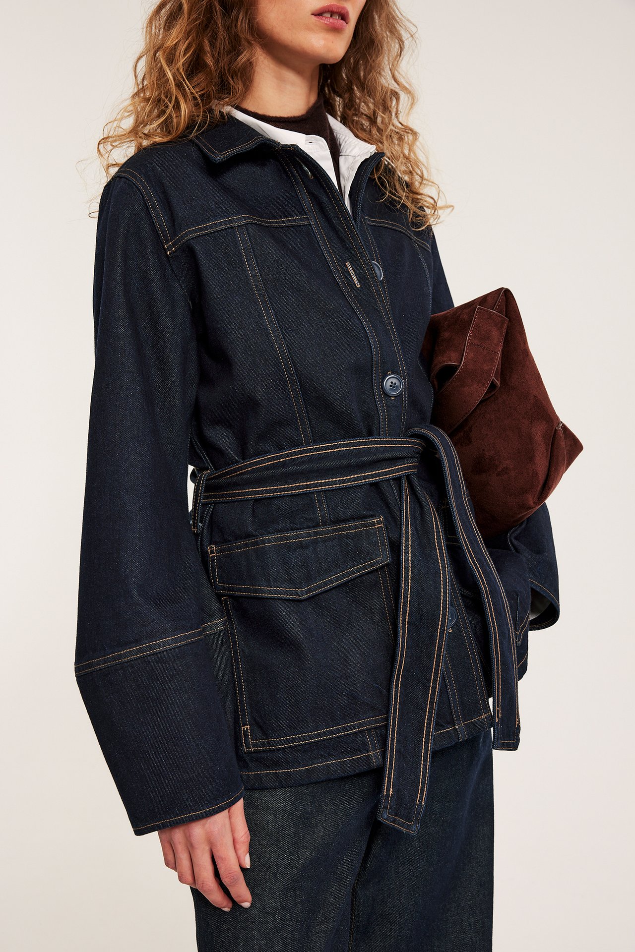 Edestä: Nainen istuvassa tummassa farkkutakissa vyöllä, valkoisen paidan päällä, pitäen ruskeaa mokkalaukkua.