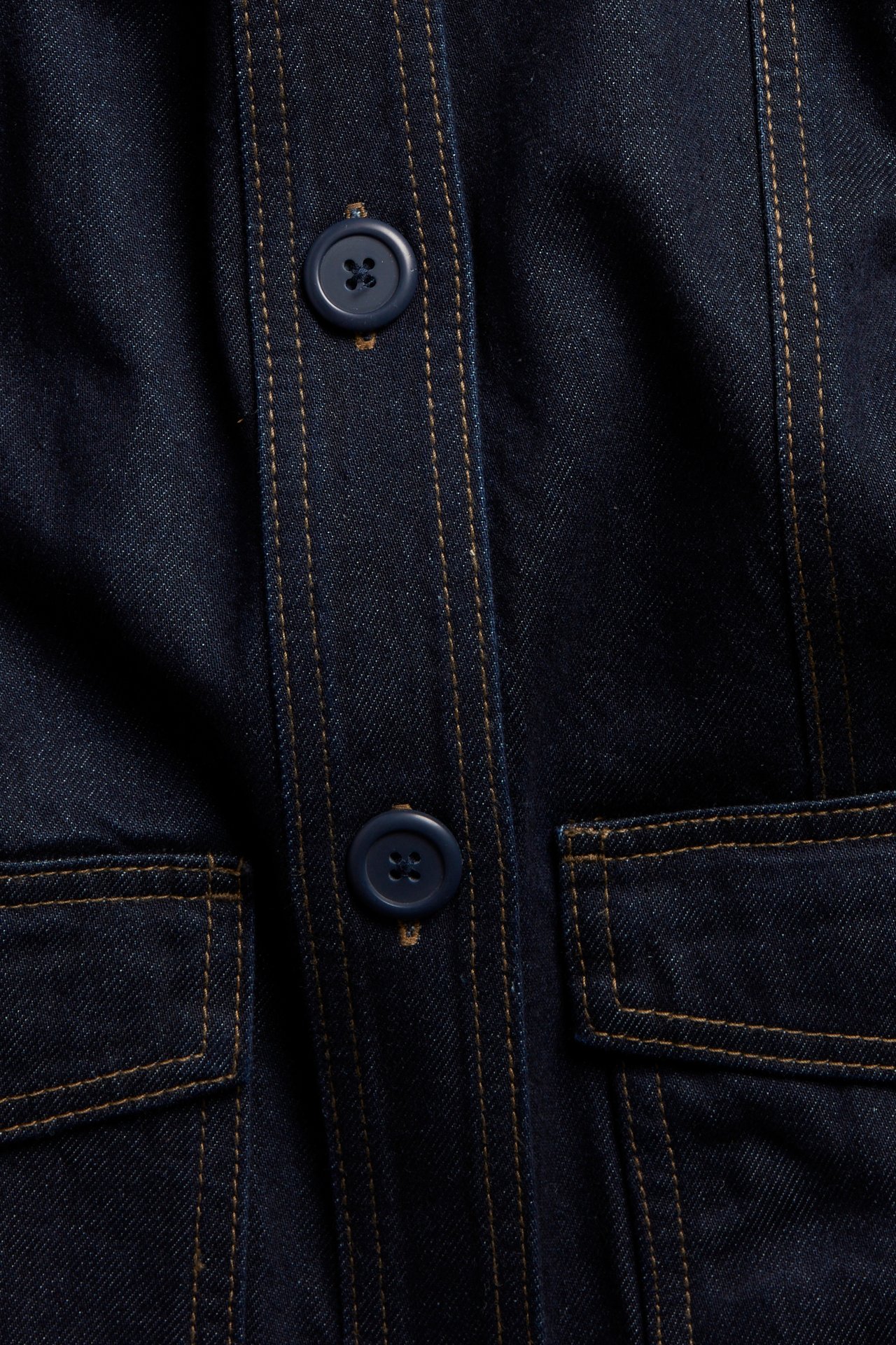 Lähikuva tummansinisestä farkkutakista ruskeilla tikkauksilla, sinisillä napeilla ja läppätaskuilla.