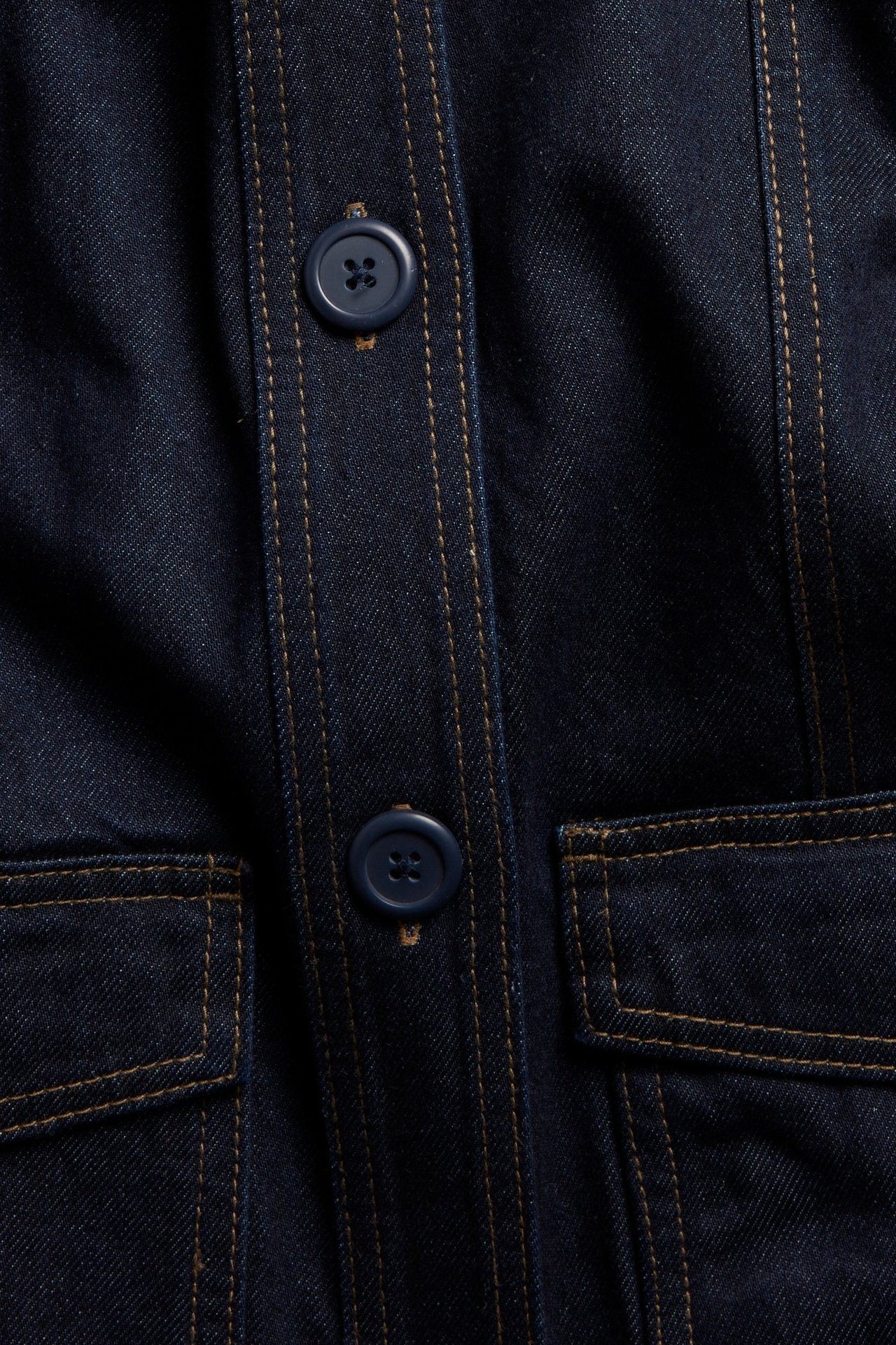 Zbliżenie na ciemnoniebieską kurtkę dżinsową z brązowymi przeszyciami, niebieskimi guzikami i patkowymi kieszeniami.