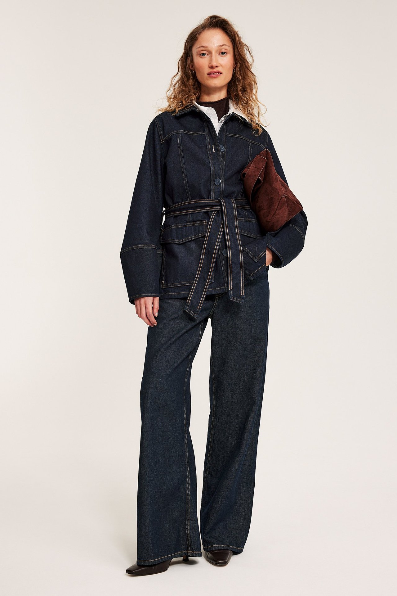 Edestä: nainen tummansinisessä vyötäröhihnaisessa farkkutakissa, yhteensopivissa leveissä farkuissa, valkoisessa paidassa, ruskea mokkanahkalaukku.