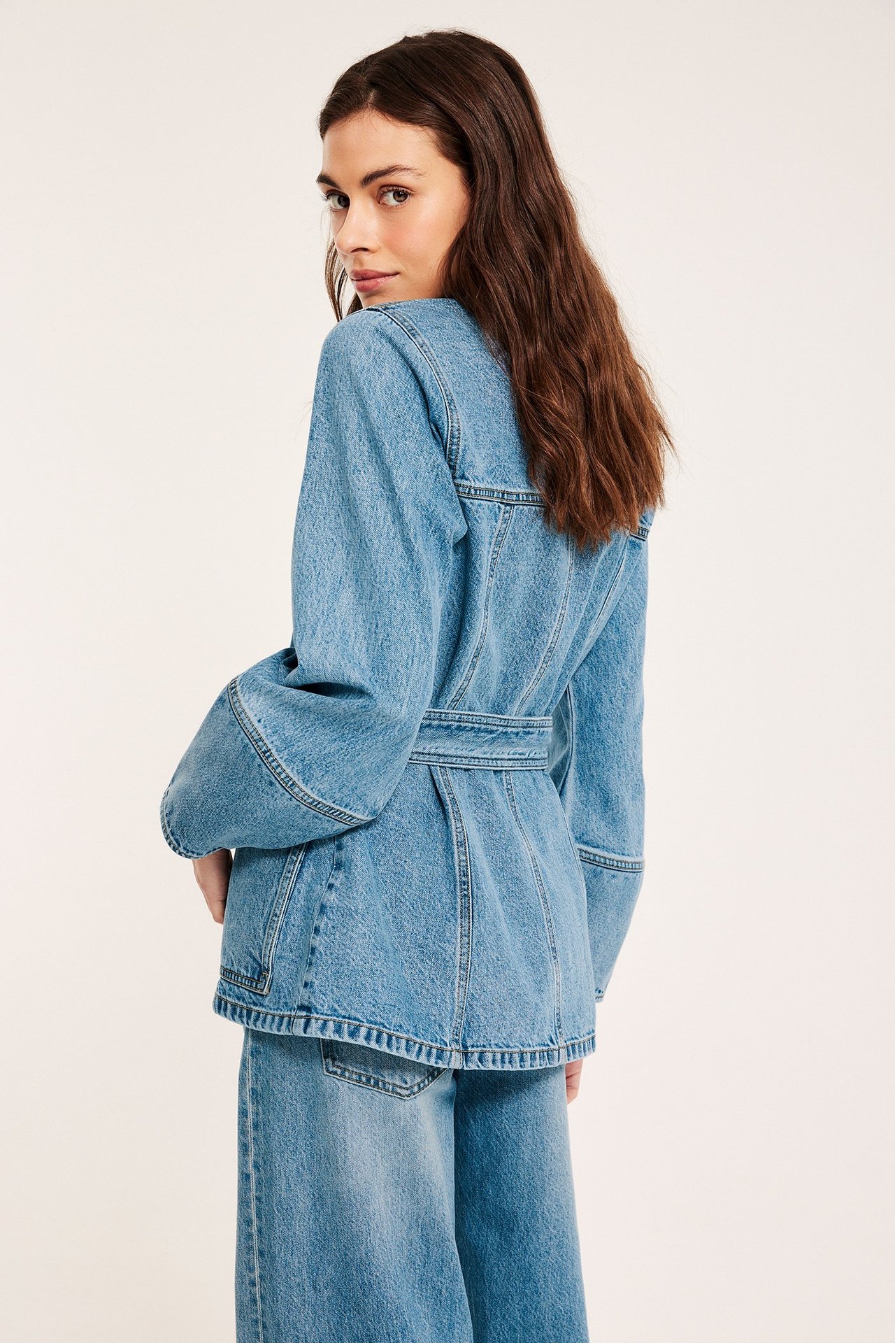 Nainen vaaleansinisessä vyötäröltä sidotussa farkkutakissa, jossa leveät hihat, ja yhteensopivissa farkkuhousuissa, takaa.