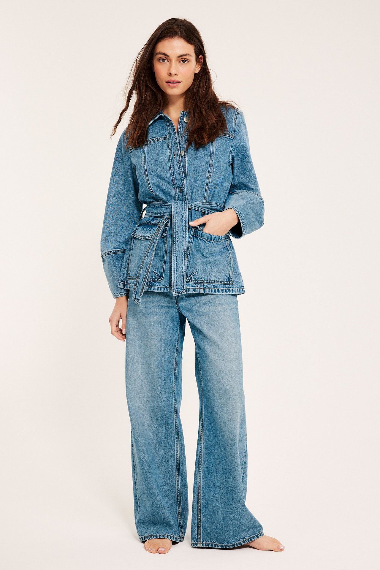 Edestä: Nainen yllään istuva vaaleansininen farkkutakki, jossa solmittu vyö, etutaskut ja leveälahkeiset farkut.