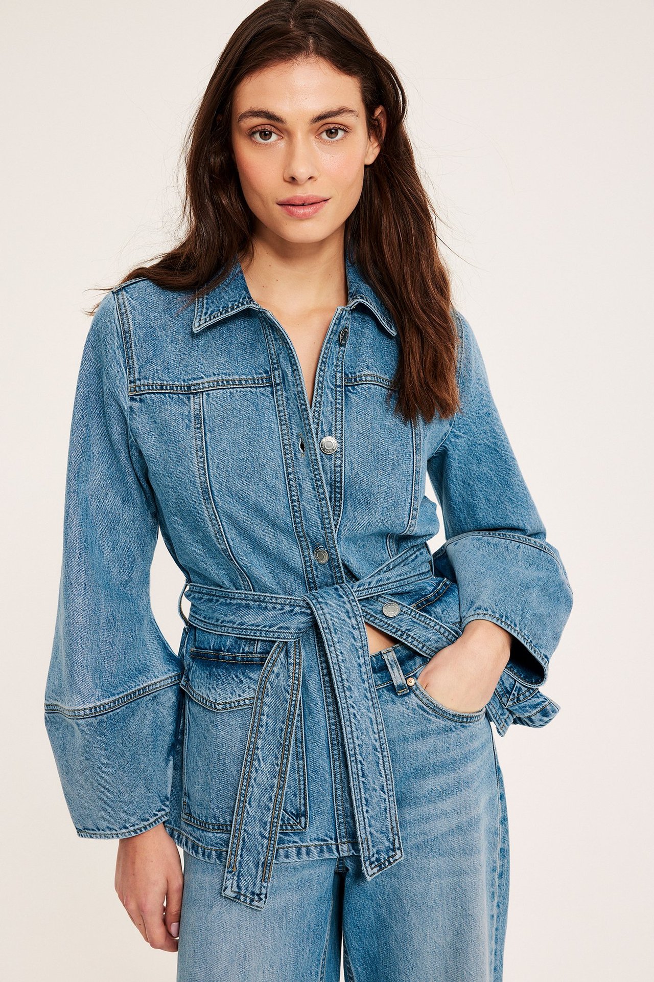 Edestä kuvattu nainen, yllään istuva farkkutakki taskuilla, sidottu vyö ja yhteensopivat farkut.