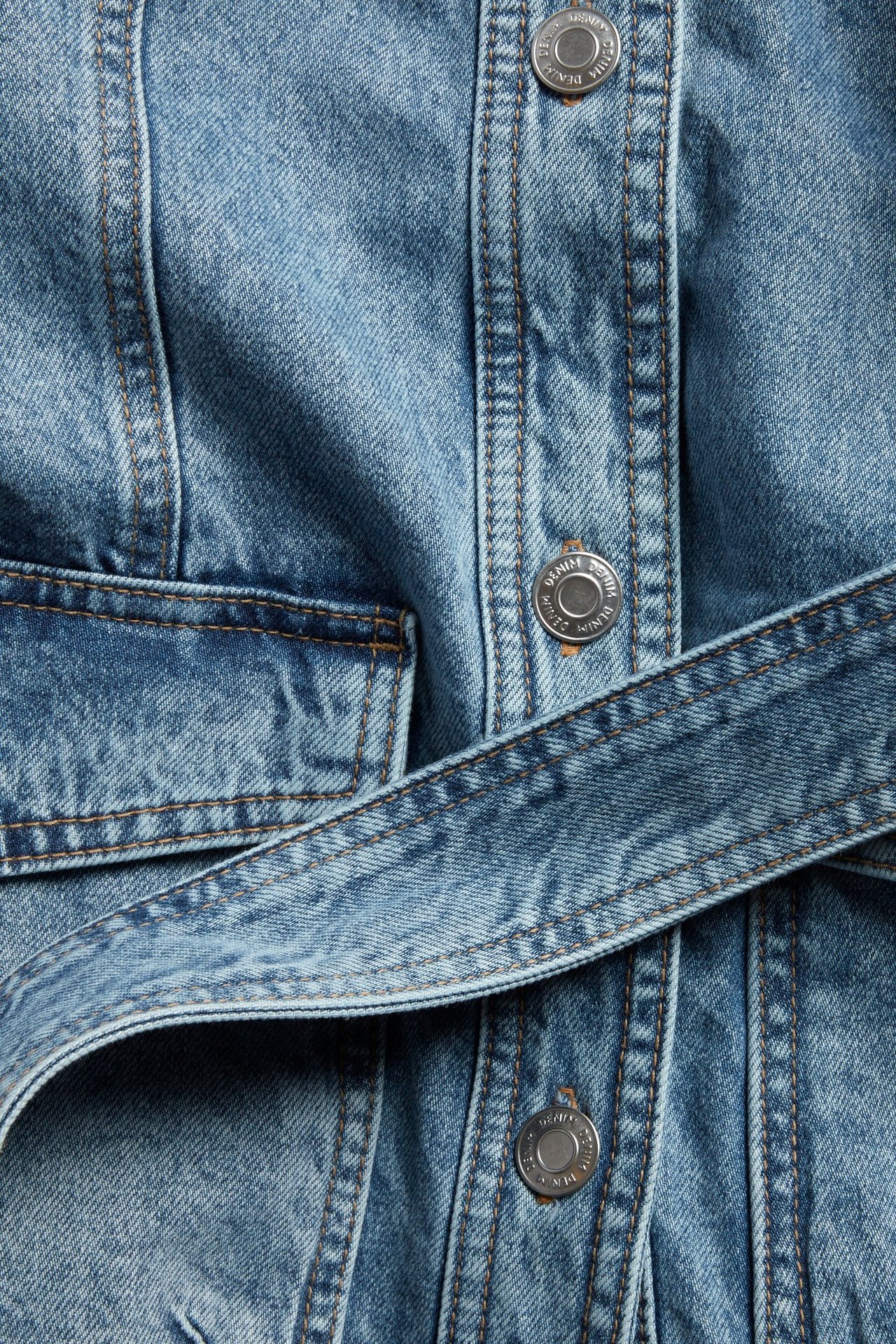 Bliski widok jasnoniebieskiej kurtki dżinsowej z metalowymi guzikami, przednią kieszenią i paskiem.