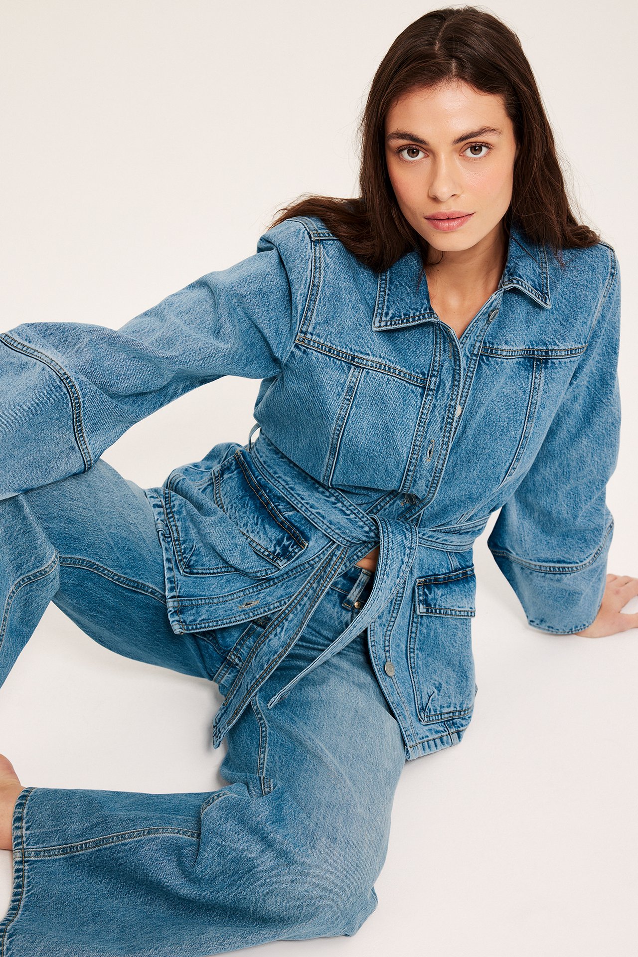 Edestäpäin: Nainen pukeutuu siniseen farkkutakkiin sidotulla vyöllä ja yhteensopiviin leveälahkeisiin farkkuhousuihin.