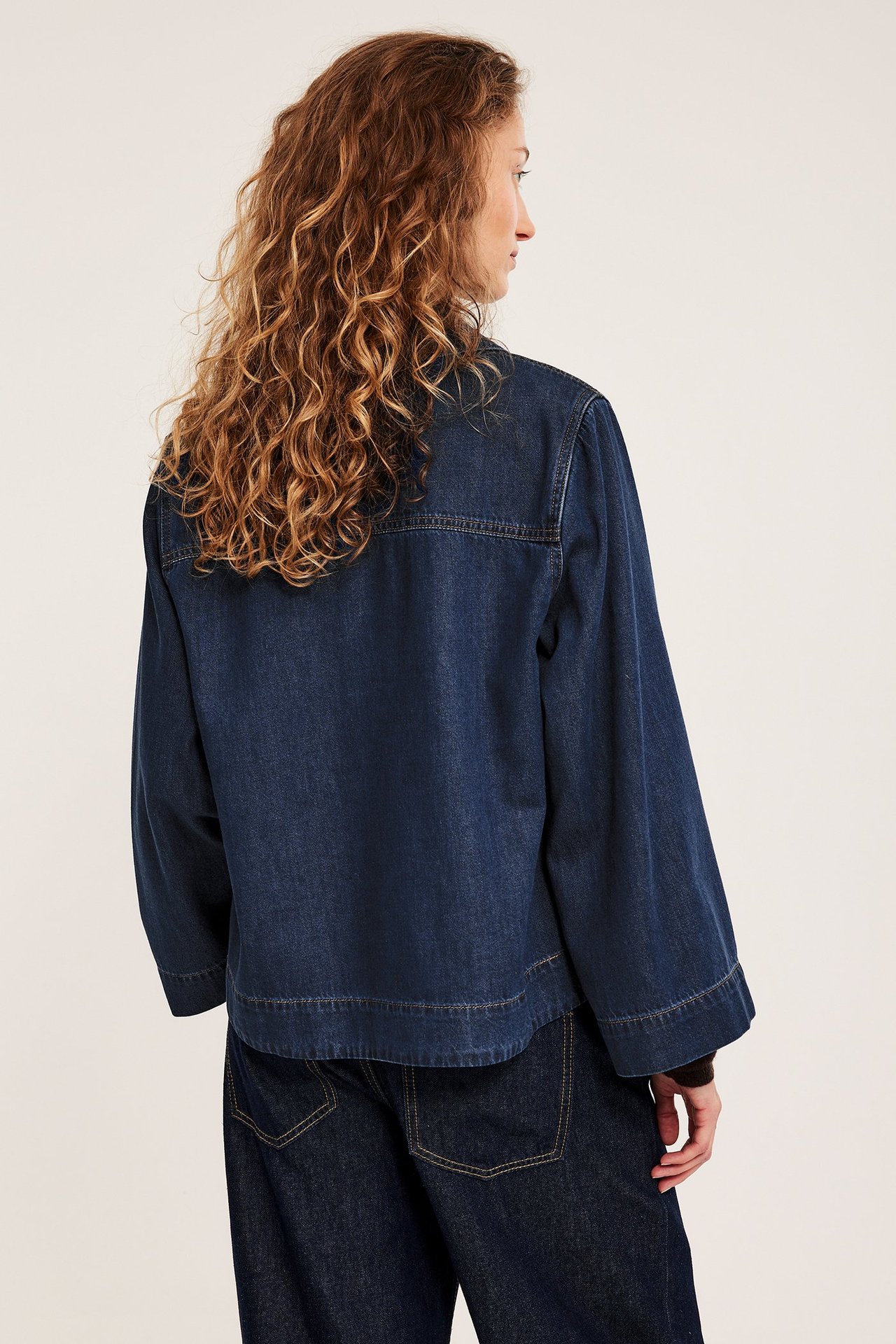 Widok z tyłu kobiety w ciemnej koszuli dżinsowej z szerokimi rękawami i ciemnych dżinsach.
