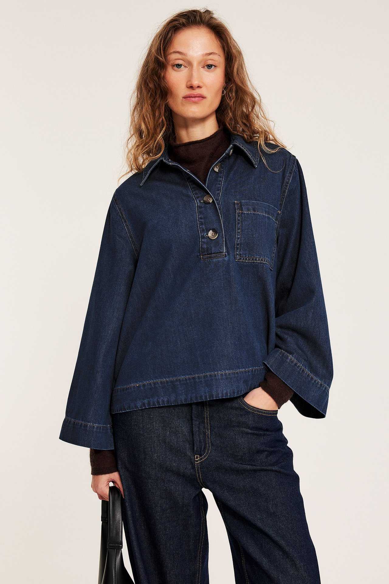 Widok z przodu: kobieta w ciemnoniebieskiej koszuli jeansowej, szerokie rękawy, ciemne jeansy, trzyma czarną torebkę.
