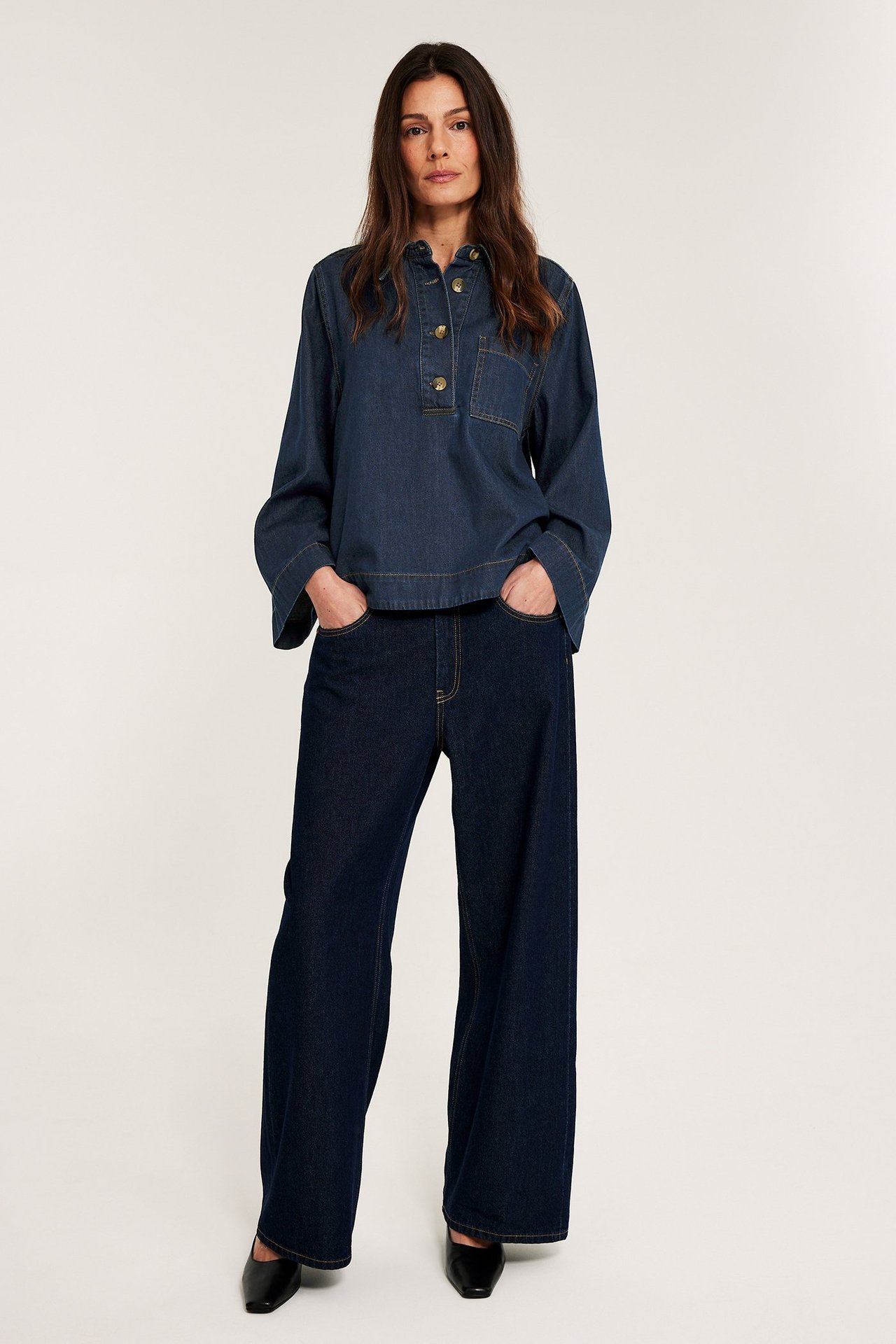 Widok z przodu, kobieta w ciemnej dżinsowej koszuli popover z guzikami imitującymi szylkret, szerokich dżinsach i czarnych butach.
