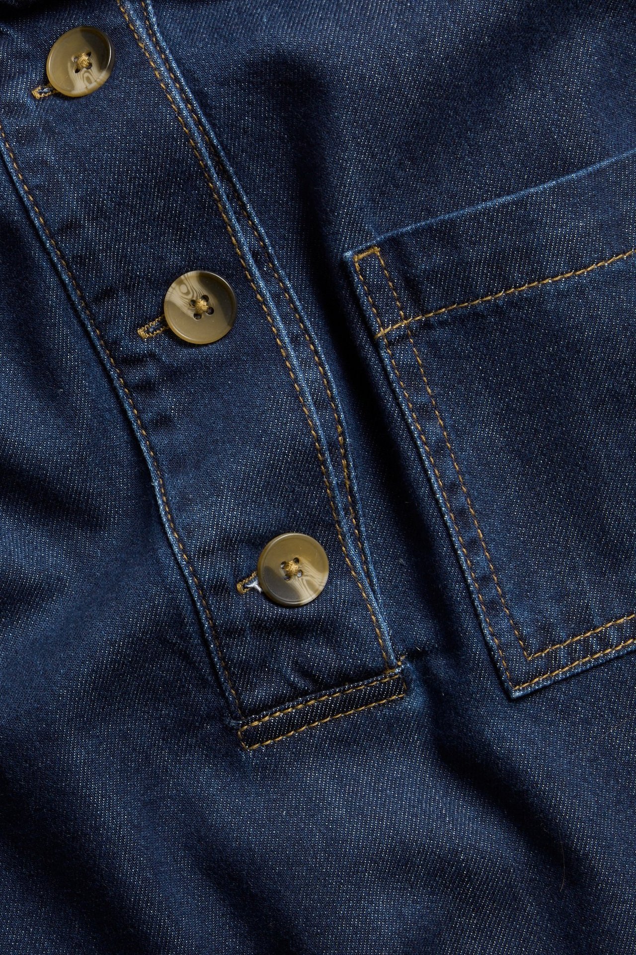 Zbliżenie ciemnej koszuli jeansowej z plisą guzikową, guzikami szylkretowymi i kieszenią na piersi.