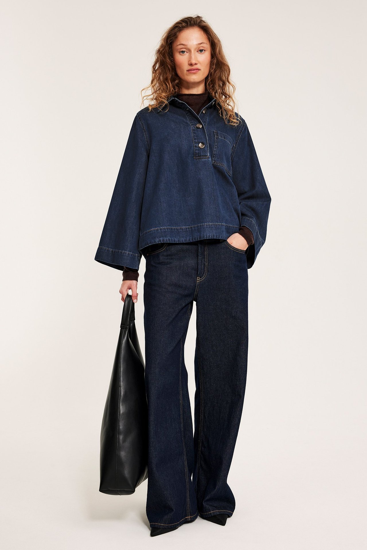 Forfra: Kvinne i mørk denimskjorte med vide ermer, mørke jeans, holder en svart veske.