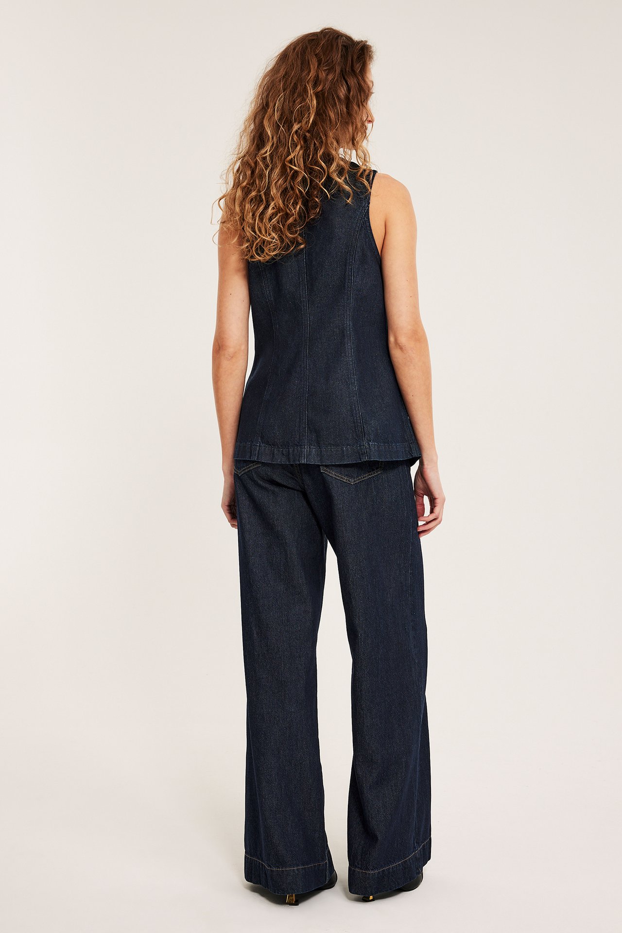 Naisen selkäkuva, yllään tummansininen istuva farkkuliivi ja tummansiniset leveälahkeiset farkut.