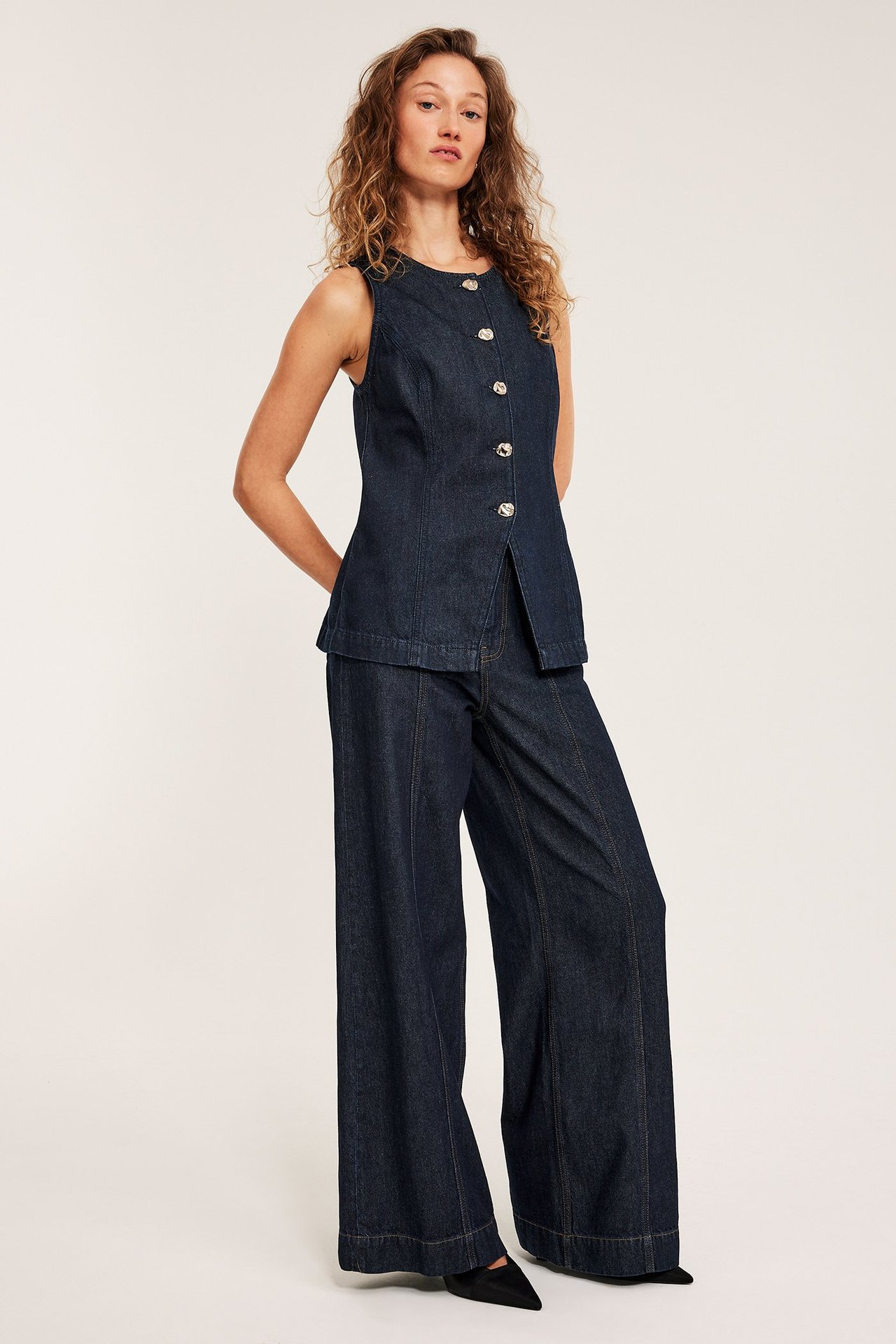 Naisen edestä: vartalonmyötäinen tummansininen farkkuliivi kultanäpeillä, yhteensopivat leveät farkut ja mustat kengät.