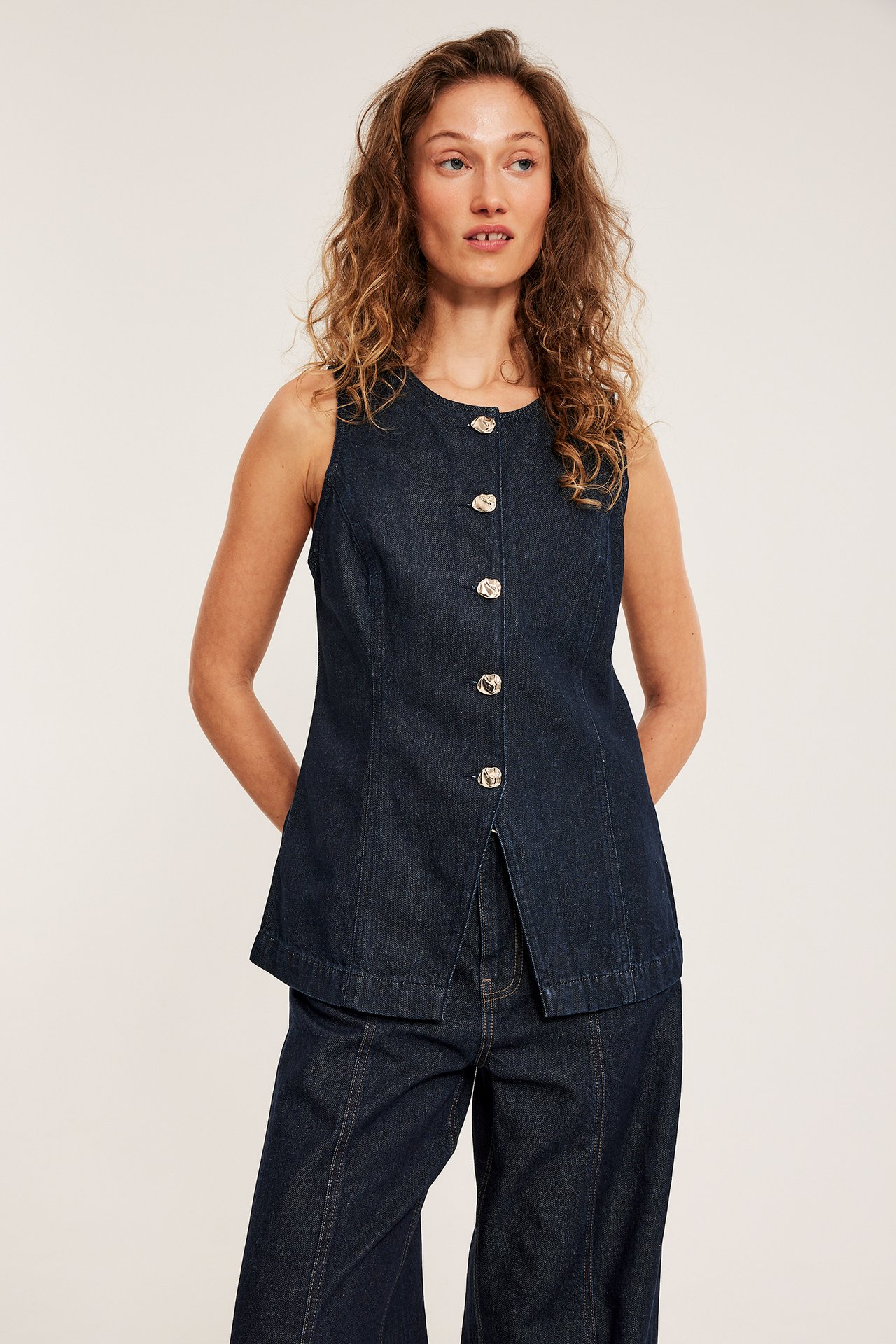 Edestä, nainen tumman farkkuvärisessä, istuvassa liivissä viidellä metallinapilla ja yhteensopivissa farkuissa.