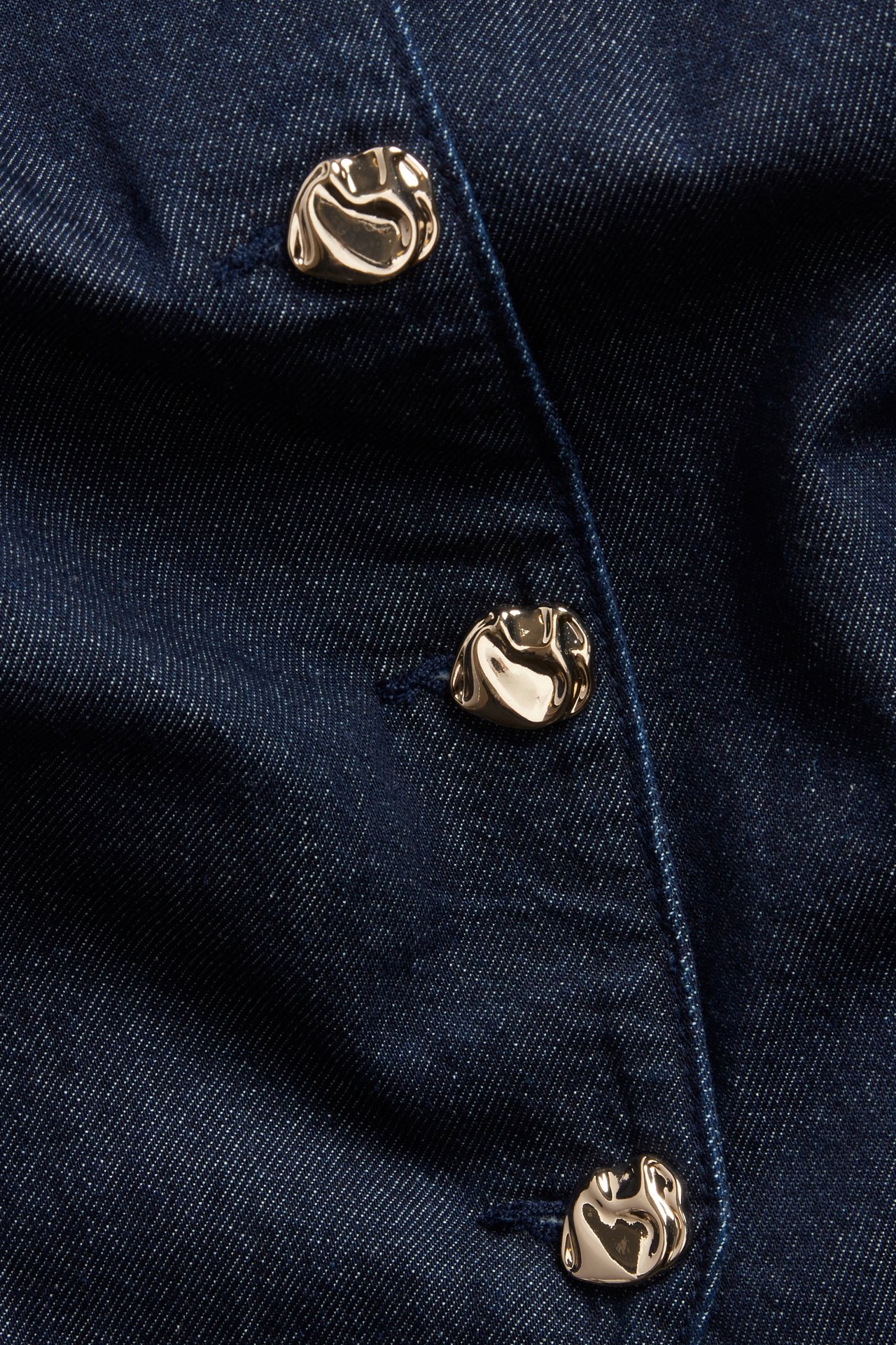 Lähikuva tummasta farkkuliivistä, jossa kolme kiiltävää kultasävyistä orgaanista metallinappia.