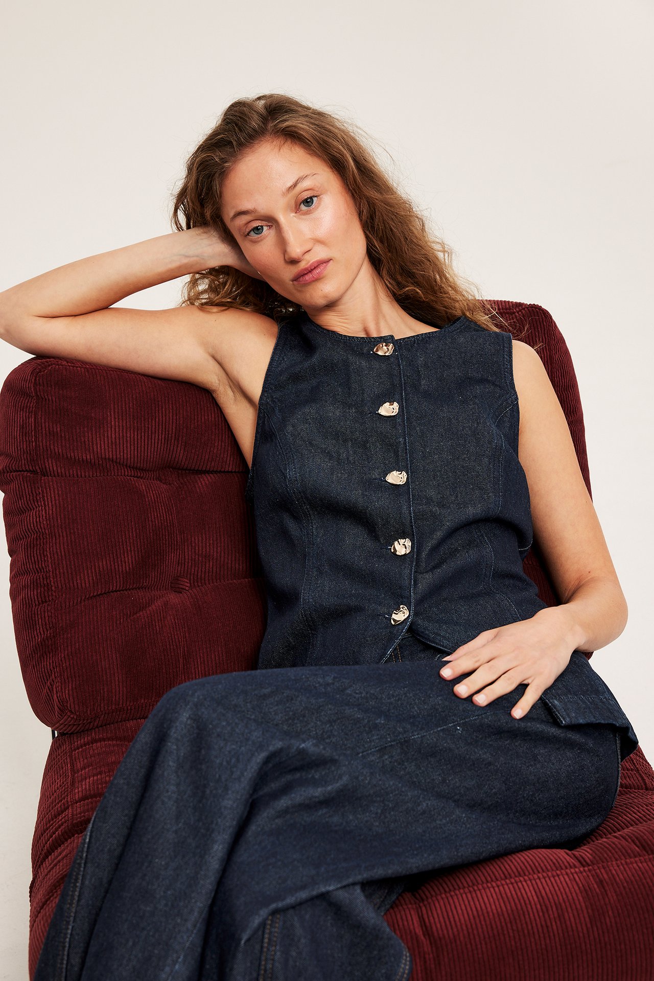 Edestä, nainen istuvassa tummassa farkkuliivissä kultanappien ja yhteensopivissa tummissa farkkuhousuissa.